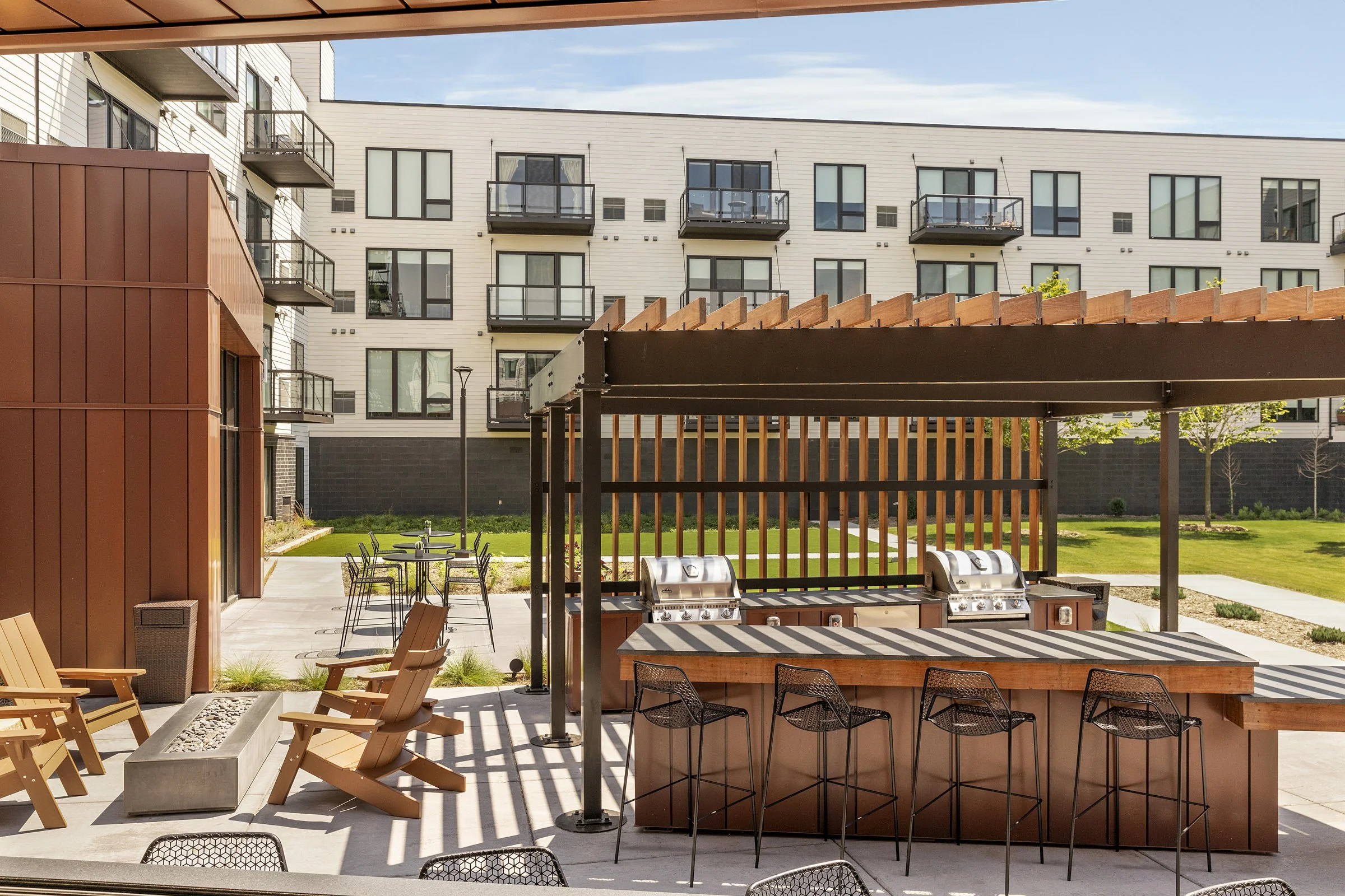 Outdoor communal area with a bar, chairs, picnic tables, some trees, and a modern apartment building in the background.