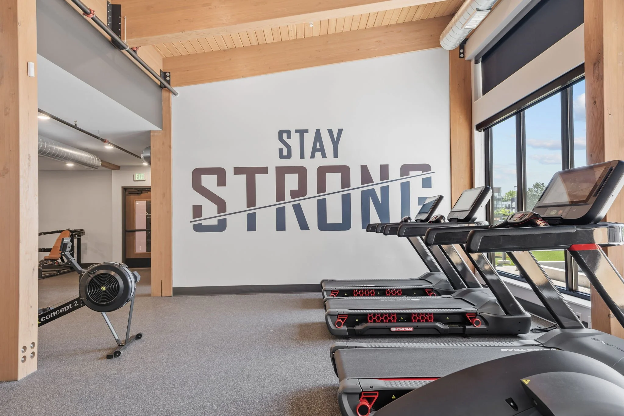 Empty gym with treadmills near large windows, wall decal says 'Stay Strong' with 'Strong' crossed out.