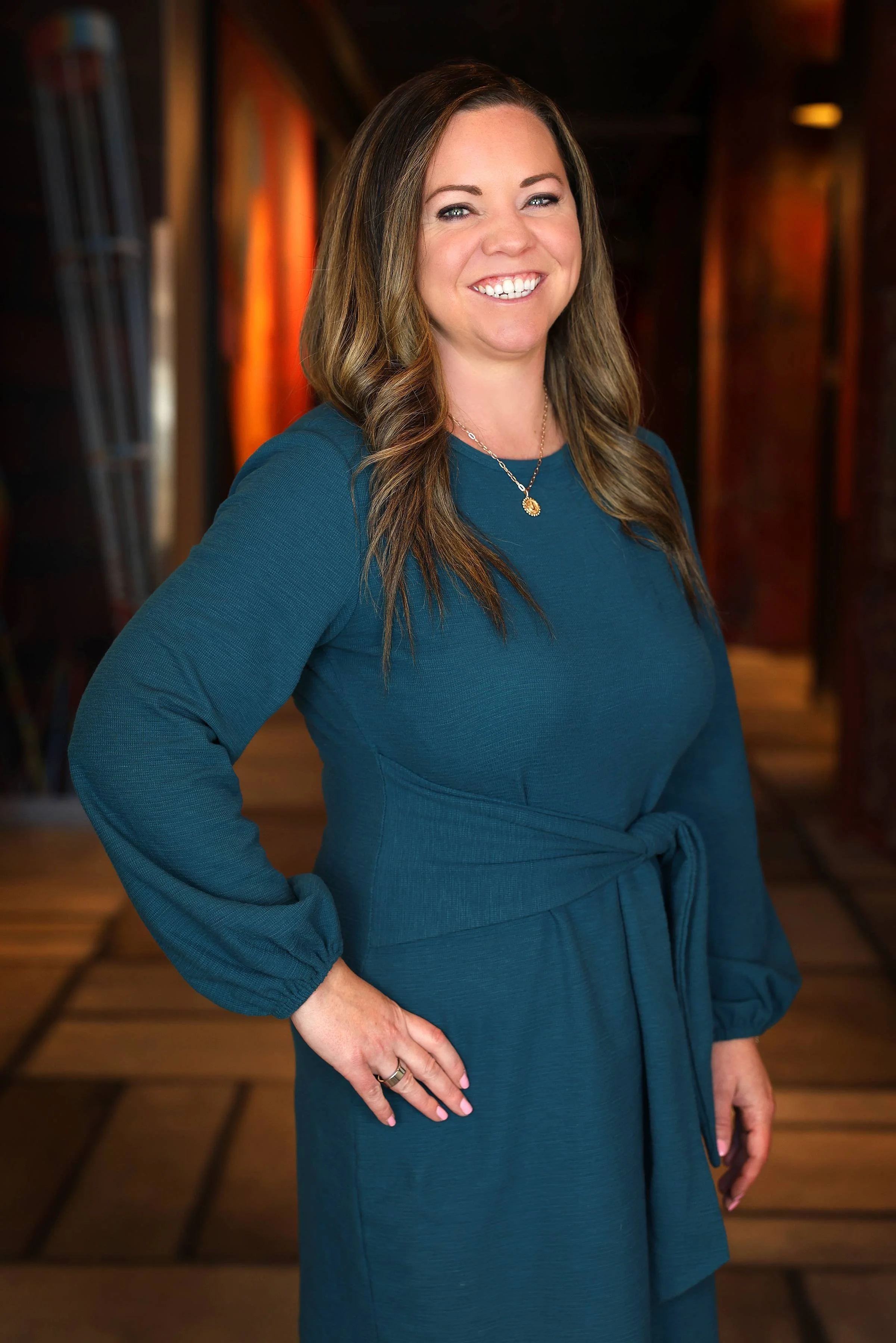 A woman with light skin, brown hair, and a bright smile, standing indoors with a warm wooden background. She is wearing a teal dress with long sleeves, a tie at the waist, a necklace, and a ring on her left hand.