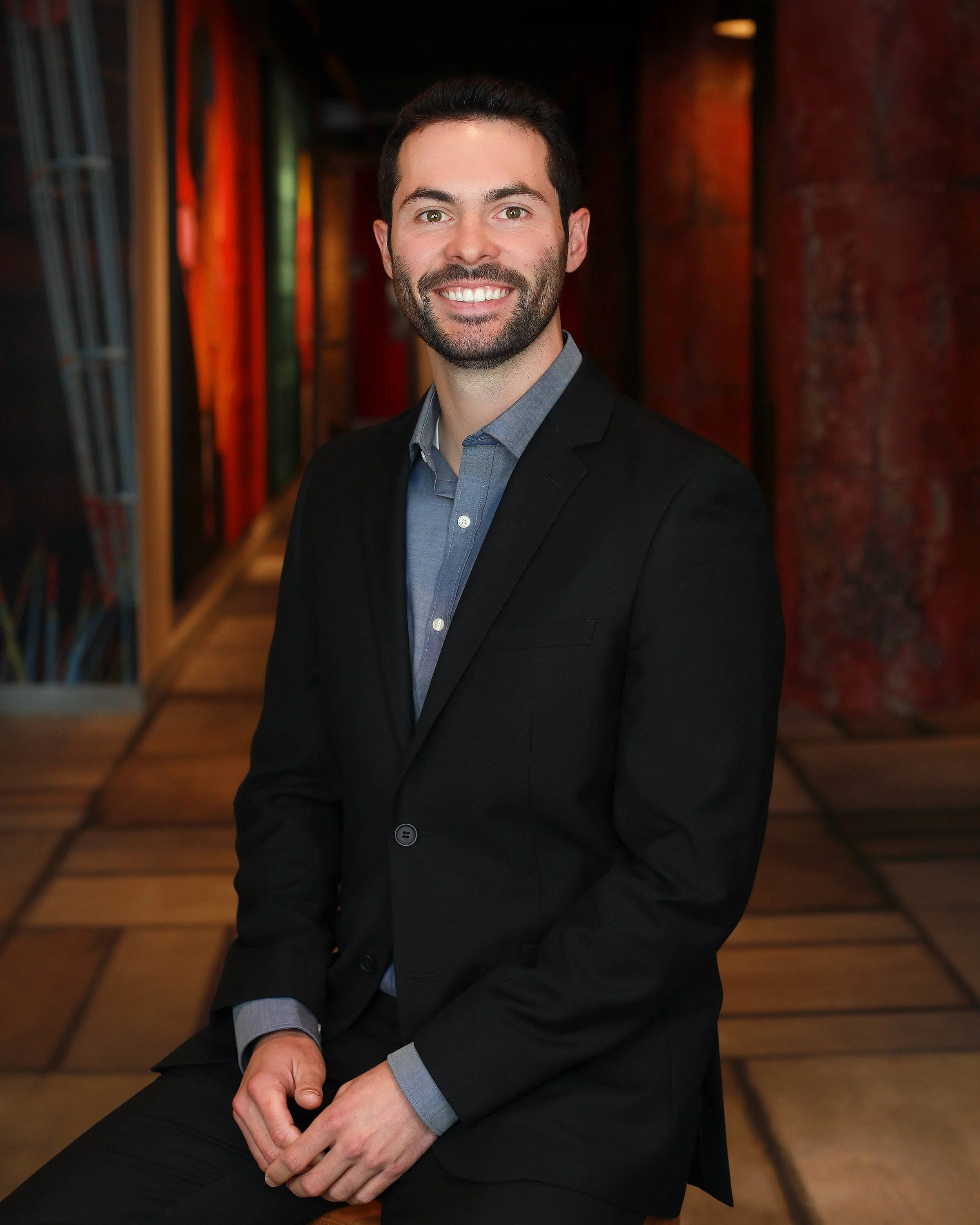 A man with dark hair and a beard, wearing a black suit and blue shirt, smiling and sitting in a corridor with colorful artwork on the walls and wooden flooring.