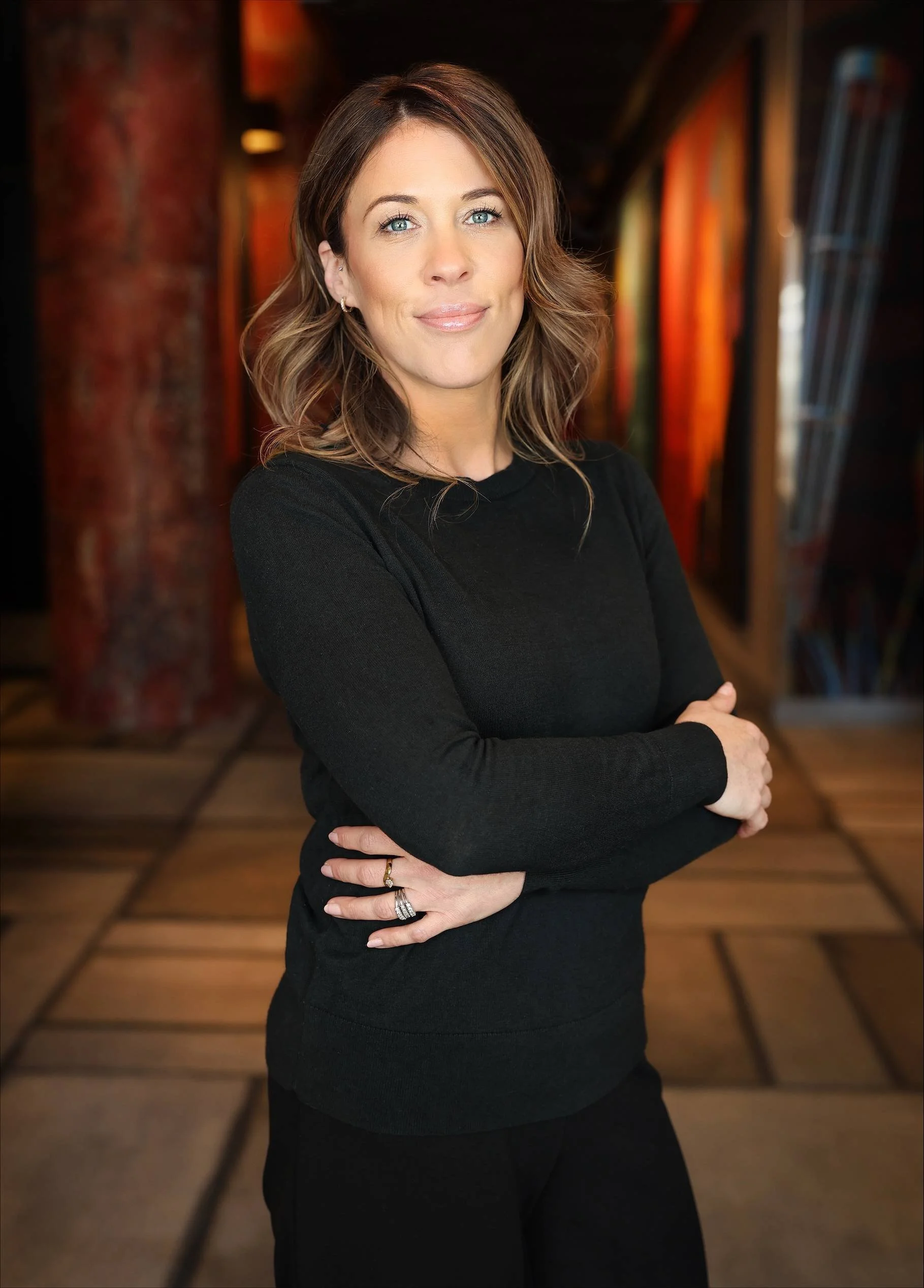 A woman with shoulder-length wavy brown hair and blue eyes standing with arms crossed in a dimly lit space with wooden floors and colorful artwork on the wall.