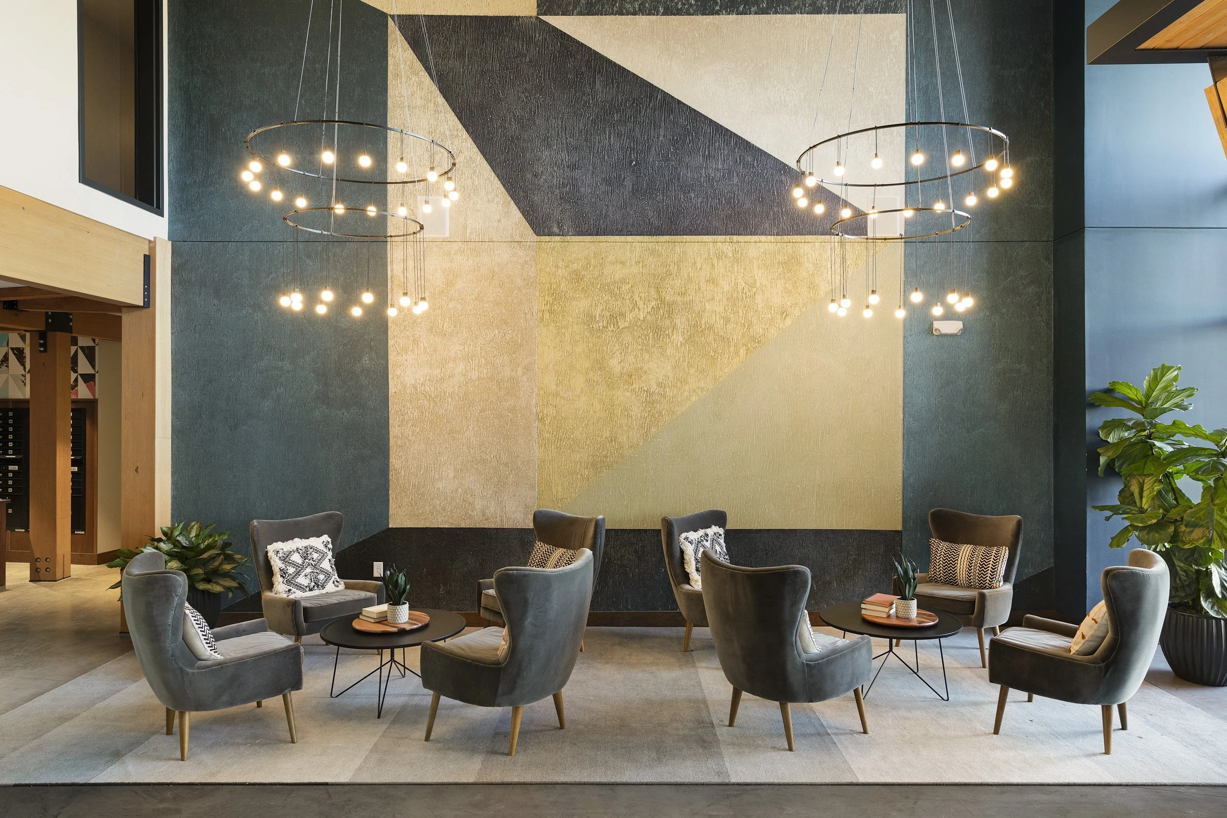 Modern seating area with six gray armchairs, round black coffee tables, and geometric wall art in shades of black, gray, beige, and gold, accented by hanging circular light fixtures and potted plants.