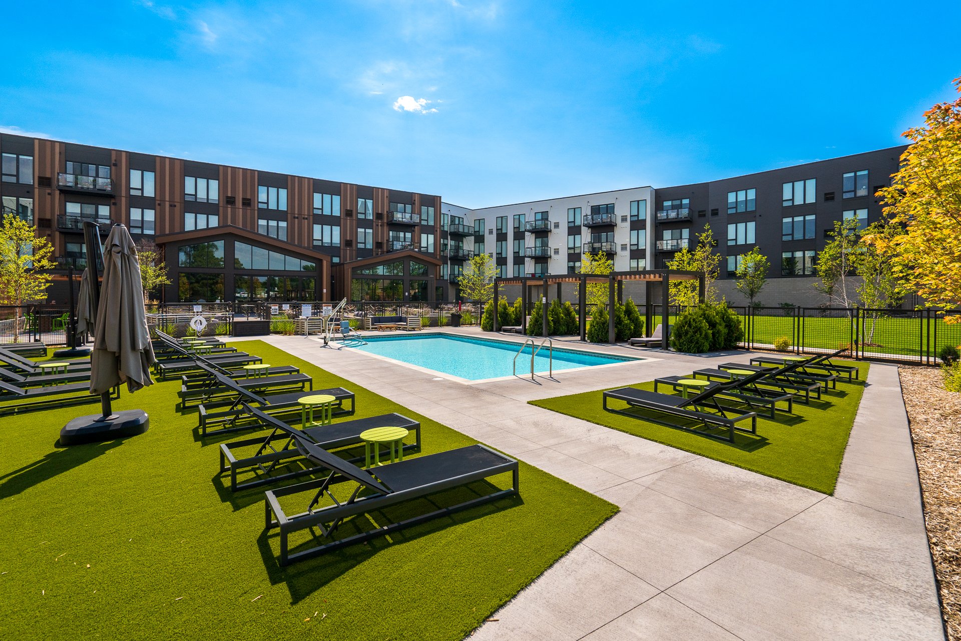 Outdoor apartment pool area with lounge chairs and umbrellas, surrounded by green grass and trees, with a modern residential building in the background under a blue sky.