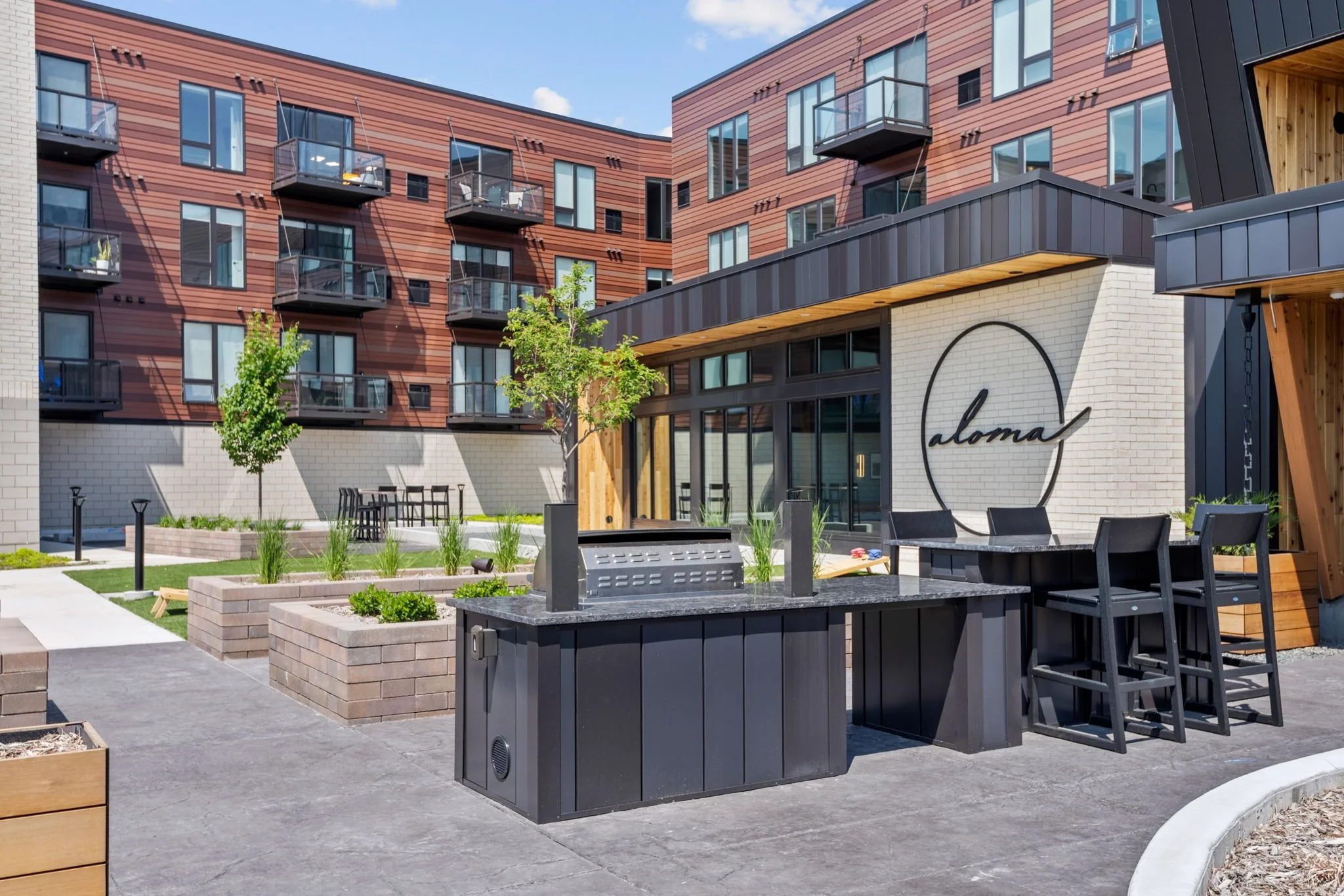 Outdoor patio with black barbecue grill, black chairs, and a table in front of a modern apartment building with balconies and a sign reading 'aloma'.