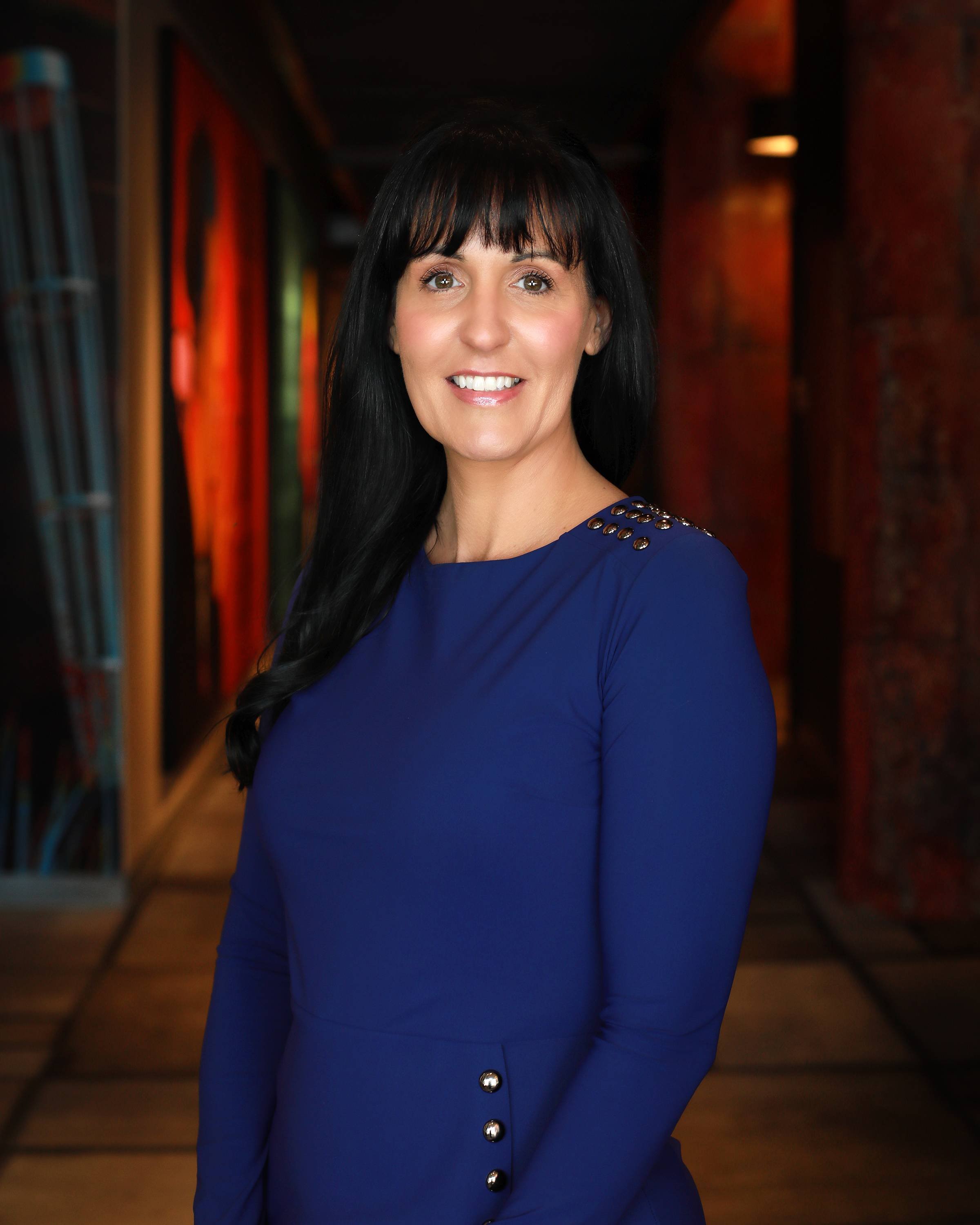 A woman with long black hair, wearing a royal blue dress with metallic button details on the shoulder and side, standing in a dimly lit, colorful background that appears to be an art gallery or museum.