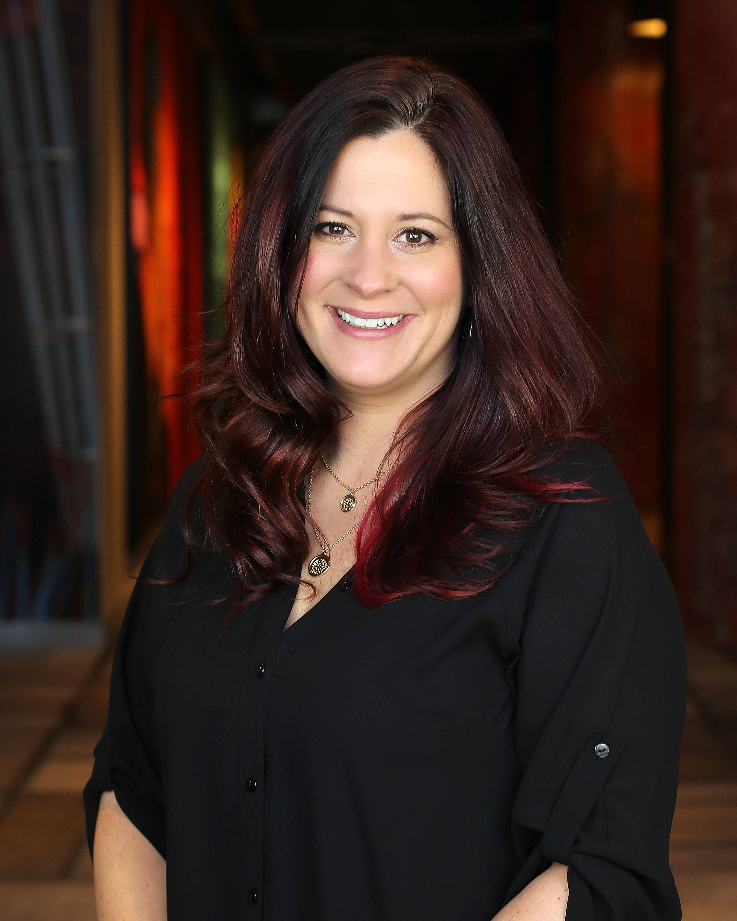 Portrait of a smiling woman with dark, burgundy hair, wearing a black blouse and layered necklaces, standing indoors with a blurred colorful background.