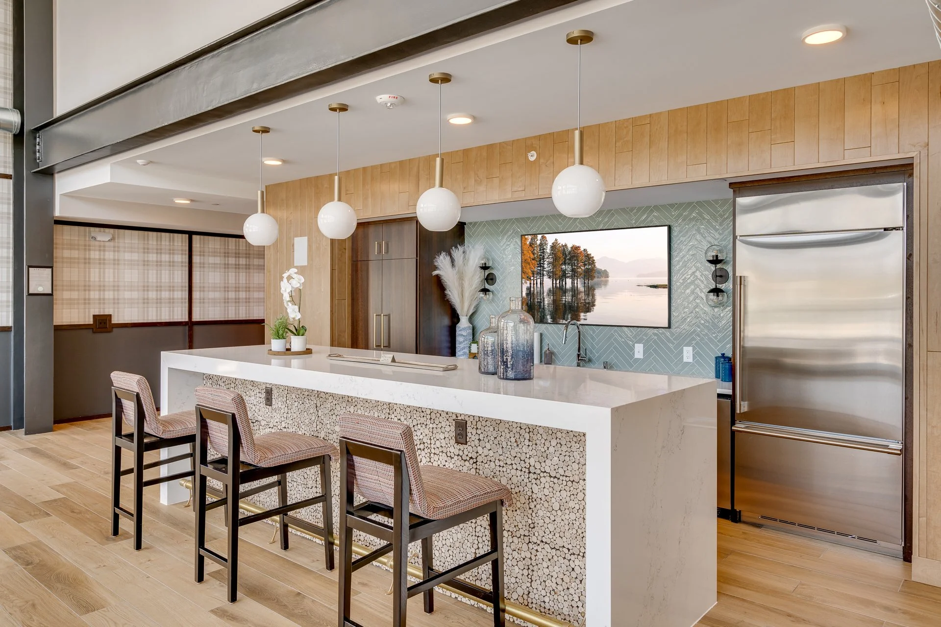Modern kitchen with a white island counter, four bar stools, a large built-in refrigerator, pendant lights, a wall-mounted television, decorative vases, and wood paneling accents.
