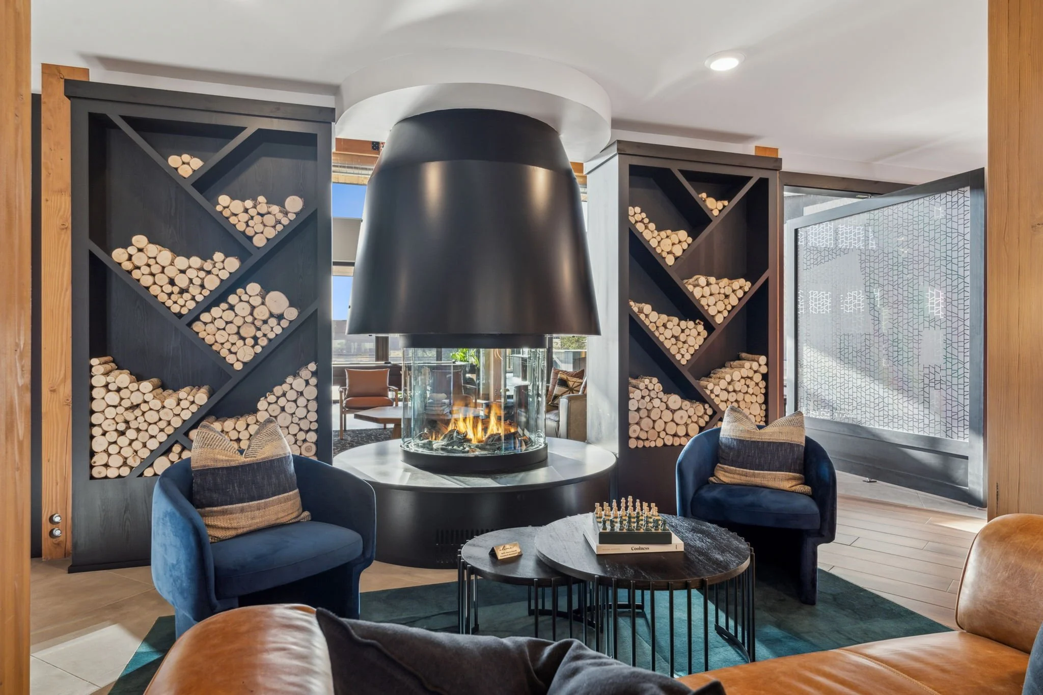 Living room with wood storage, fireplace, blue chairs, round coffee table with chessboard, leather sofa, and window with geometric patterned glass.