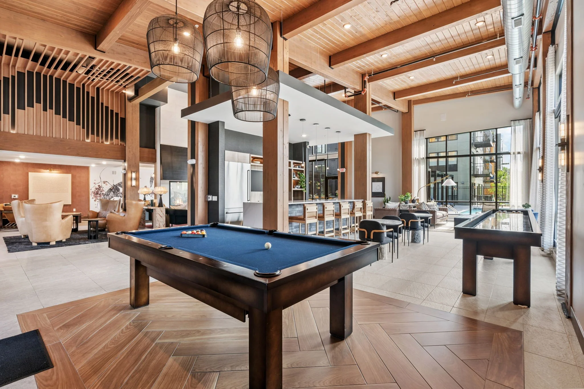 Interior of a modern lounge with a pool table, comfortable chairs, bar stools, and large windows with natural light.