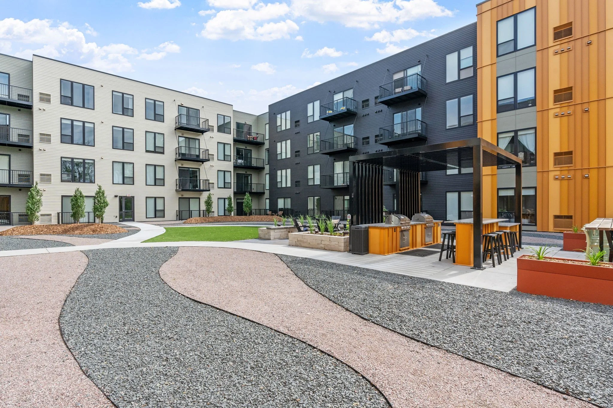 View of an outdoor courtyard with modern apartment building, walking paths, landscaped greenery, and a covered outdoor grill area with seating.