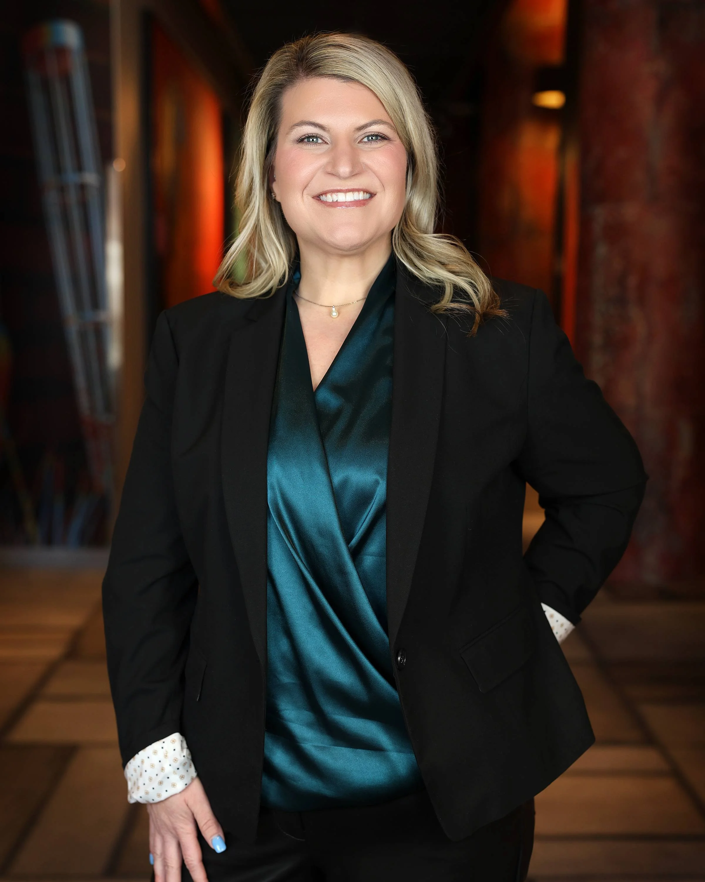 A woman with blonde hair wearing a black blazer over a teal satin blouse, standing in a warmly lit interior with wood floors and brick walls, smiling at the camera.