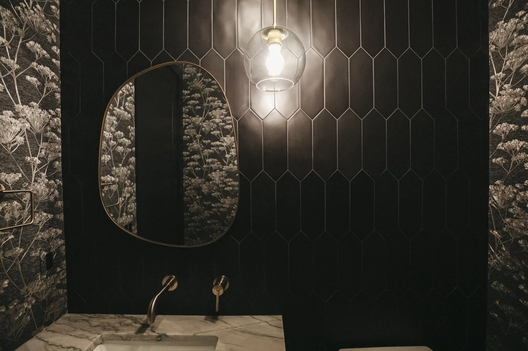 Modern bathroom with black hexagonal tile wall, floral wallpaper, oval mirror with a gold frame, marble countertop with a sink, gold faucet, and a glass pendant light above.