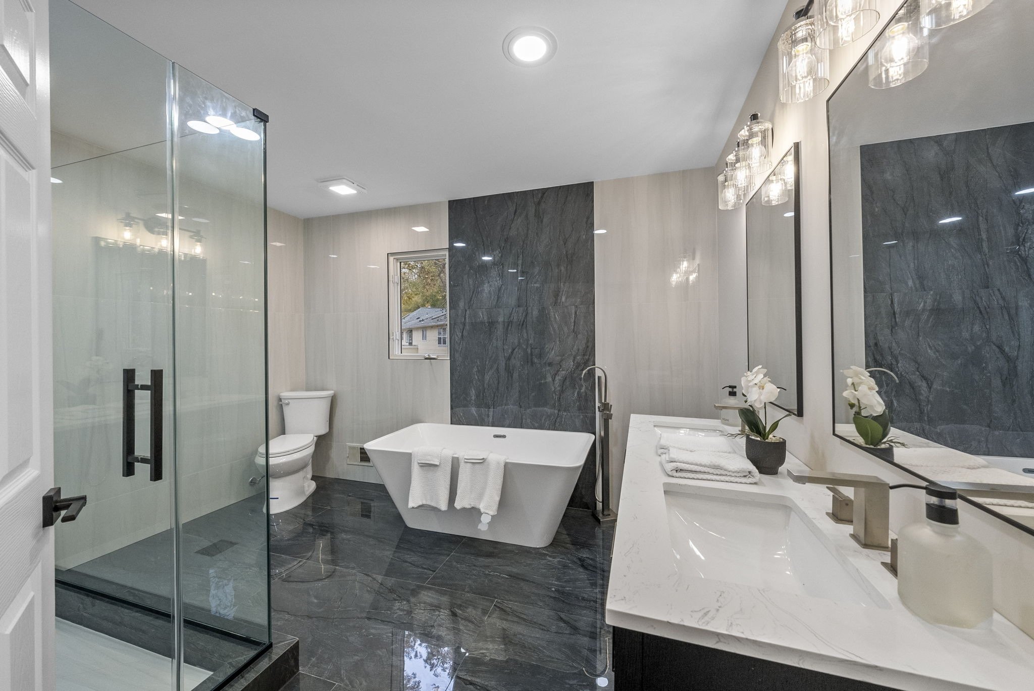 Modern bathroom with a glass-enclosed shower, a freestanding tub, a toilet, a window, a marble countertop with a sink, towels, and decorative plants.