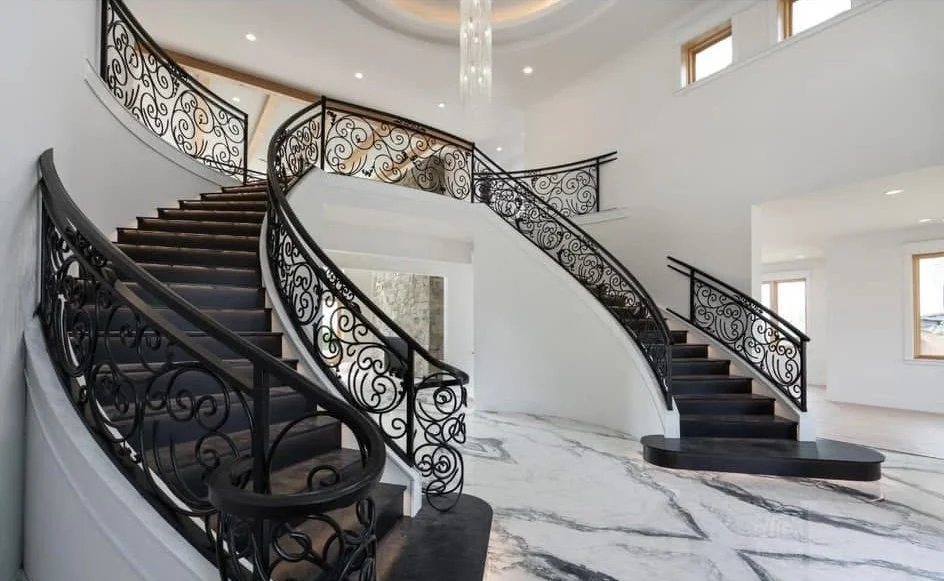 Elegant curved staircase with ornate black wrought iron railing in a modern luxury home interior featuring high ceilings and marble flooring.