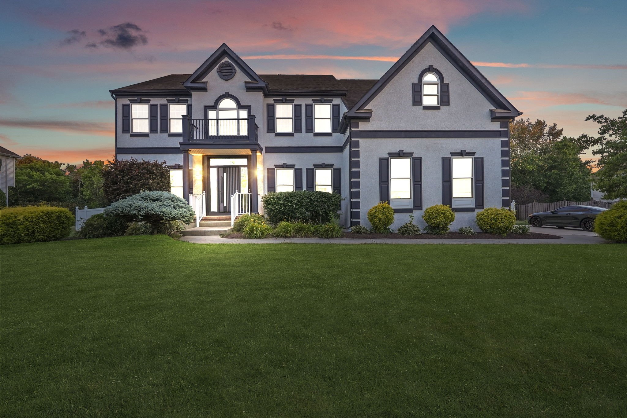 A large, two-story house with a neatly manicured lawn during sunset. The house has a white exterior with black shutters, a front porch with stairs, and a balcony on the second floor. There are bushes and flower beds in front, and a driveway with a bl