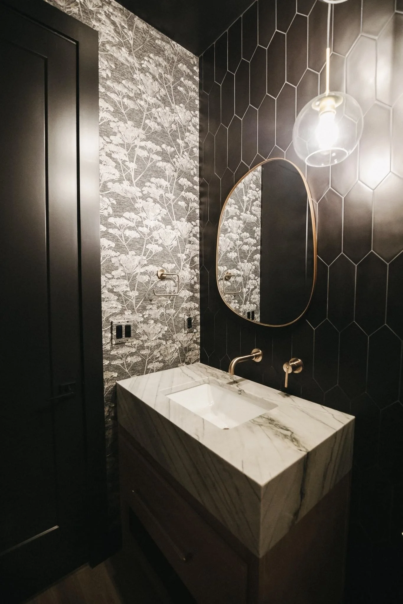 A modern bathroom vanity with a rectangular marble sink, a round mirror with a gold frame, black hexagonal tiled wall, floral wallpaper, and a glass pendant light.
