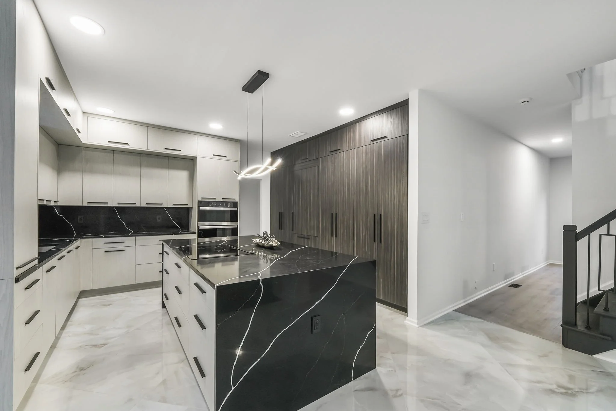 Modern kitchen with black marble island, white cabinetry, black backsplash with white veining, built-in oven, and dark wood cabinets, open space with staircase in background.