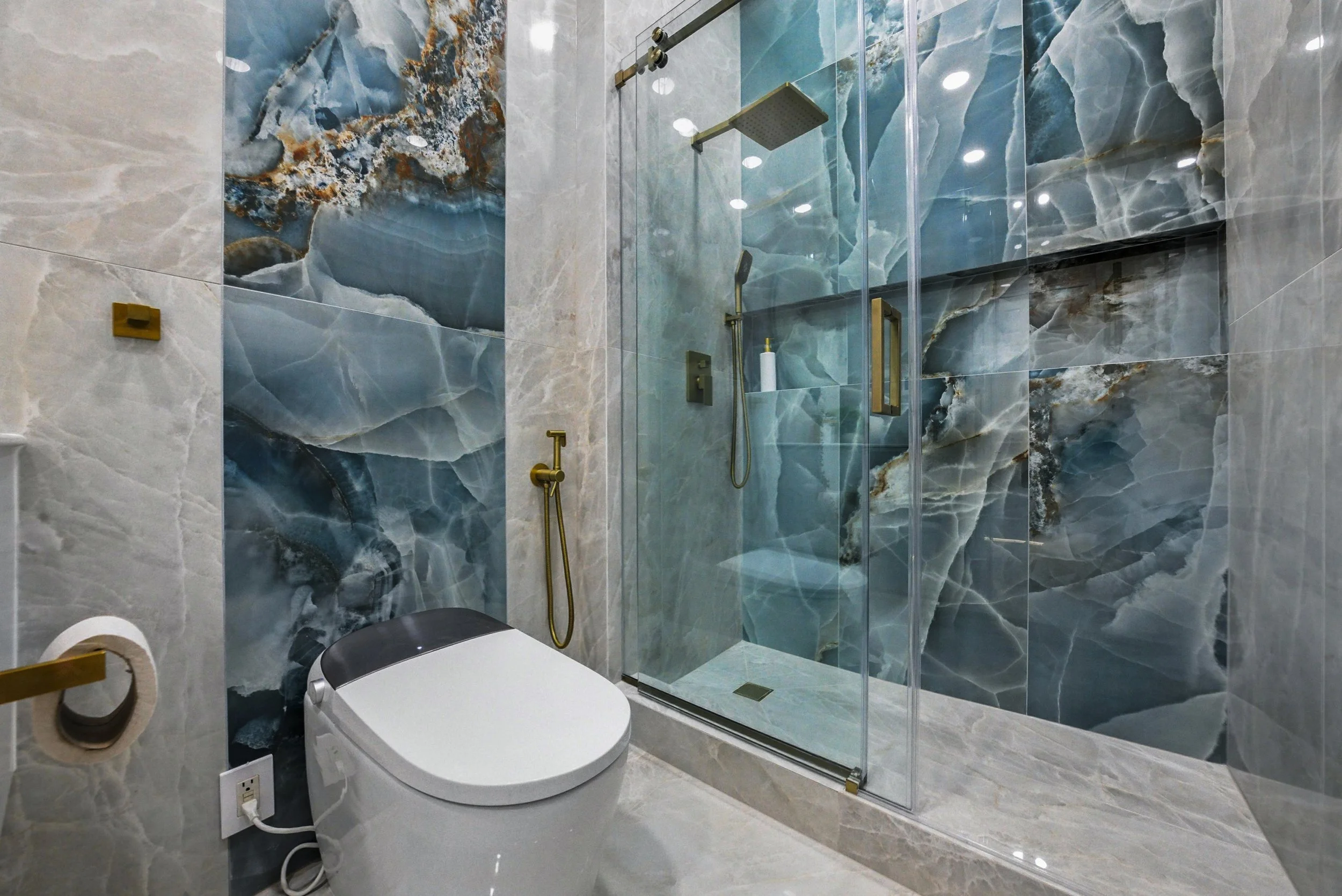 A modern bathroom featuring a wall with large polished marble tiles in blue and gray, a glass-enclosed shower with a gold handle and fixtures, and a white toilet adjacent to the shower.