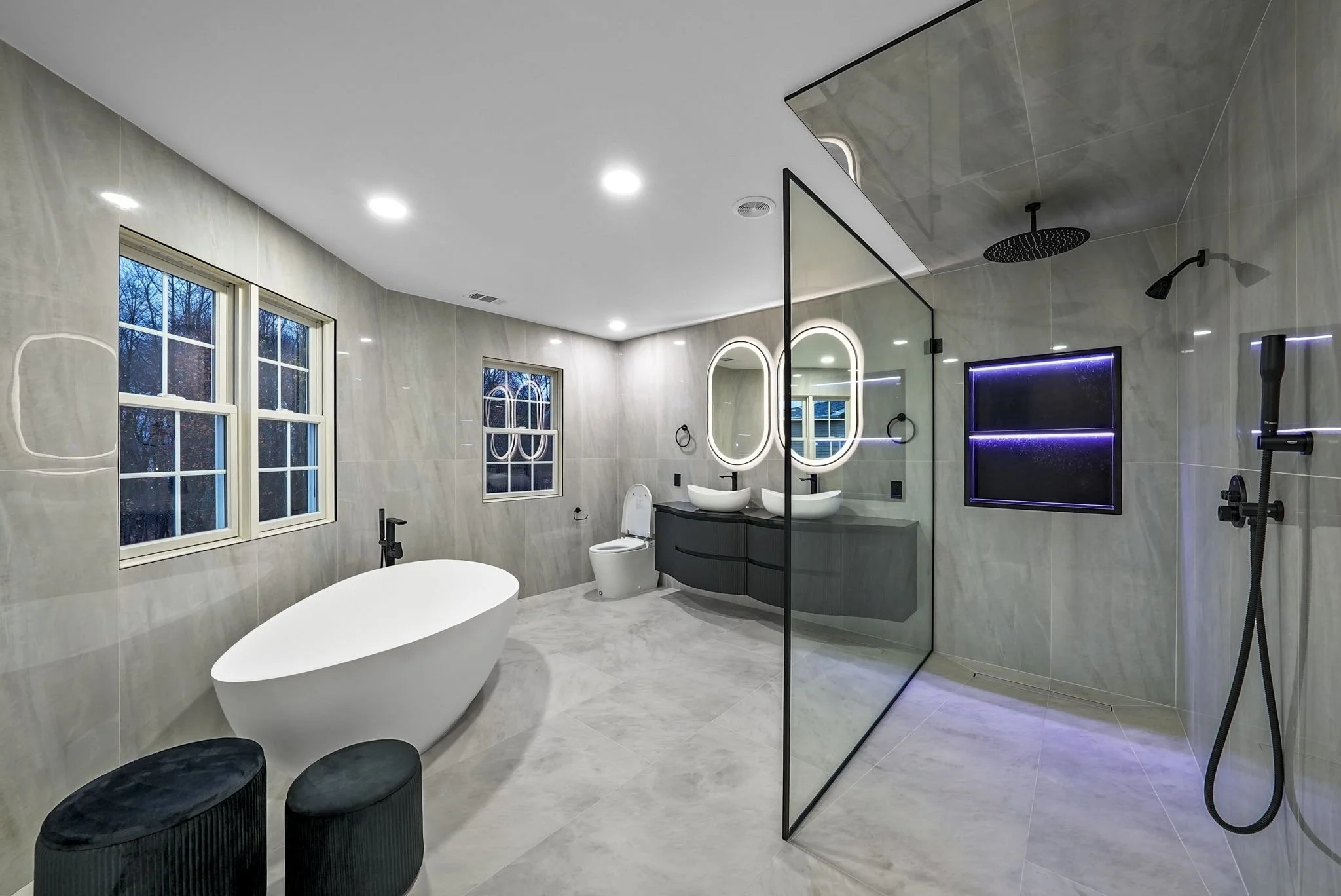 Modern bathroom with a freestanding bathtub, dual sinks with illuminated mirrors, a glass shower with a rainfall showerhead, and black accents, including window frames, faucets, and stools.