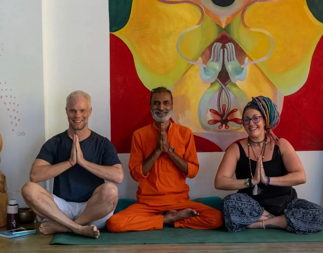Three people sitting cross-legged on a yoga mat, praying with hands in a namaste gesture, in front of a colorful abstract mural featuring a symmetrical design with floral and organic shapes.