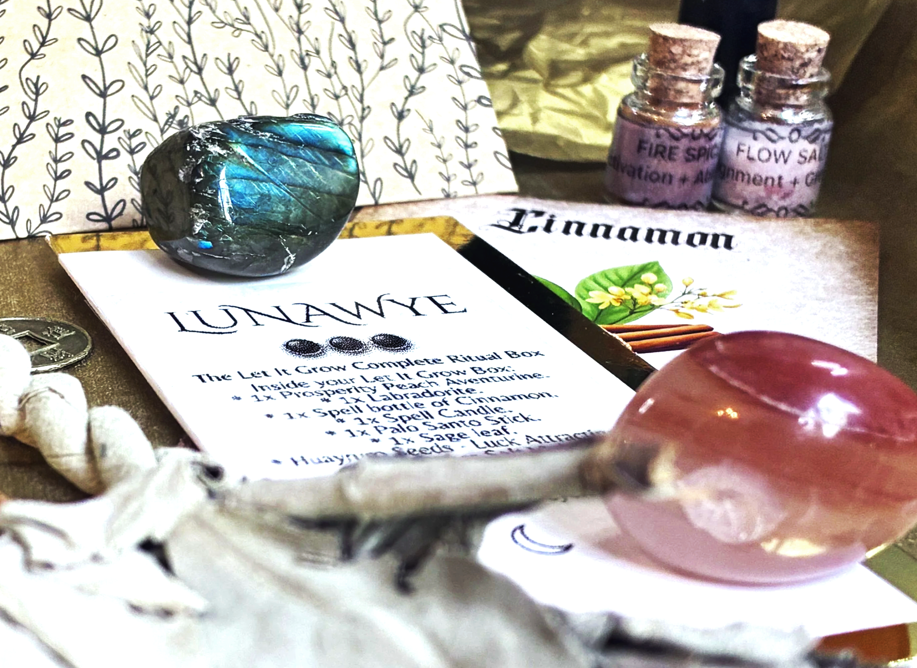 A collection of spiritual and ritual items including a labradorite stone, two small bottles labeled 'Fire Spice' and 'Flow Salt,' a pink rose quartz crystal, and a printed ritual box list titled 'LUNAWYE The Let It Grow Complete Ritual Box' with instructions and decorative elements.