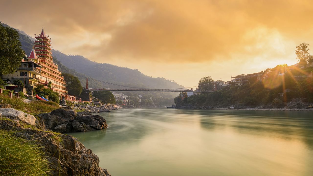 Sunset over a river with buildings and hills on either side, a bridge crossing the river, and sun rays shining through clouds.