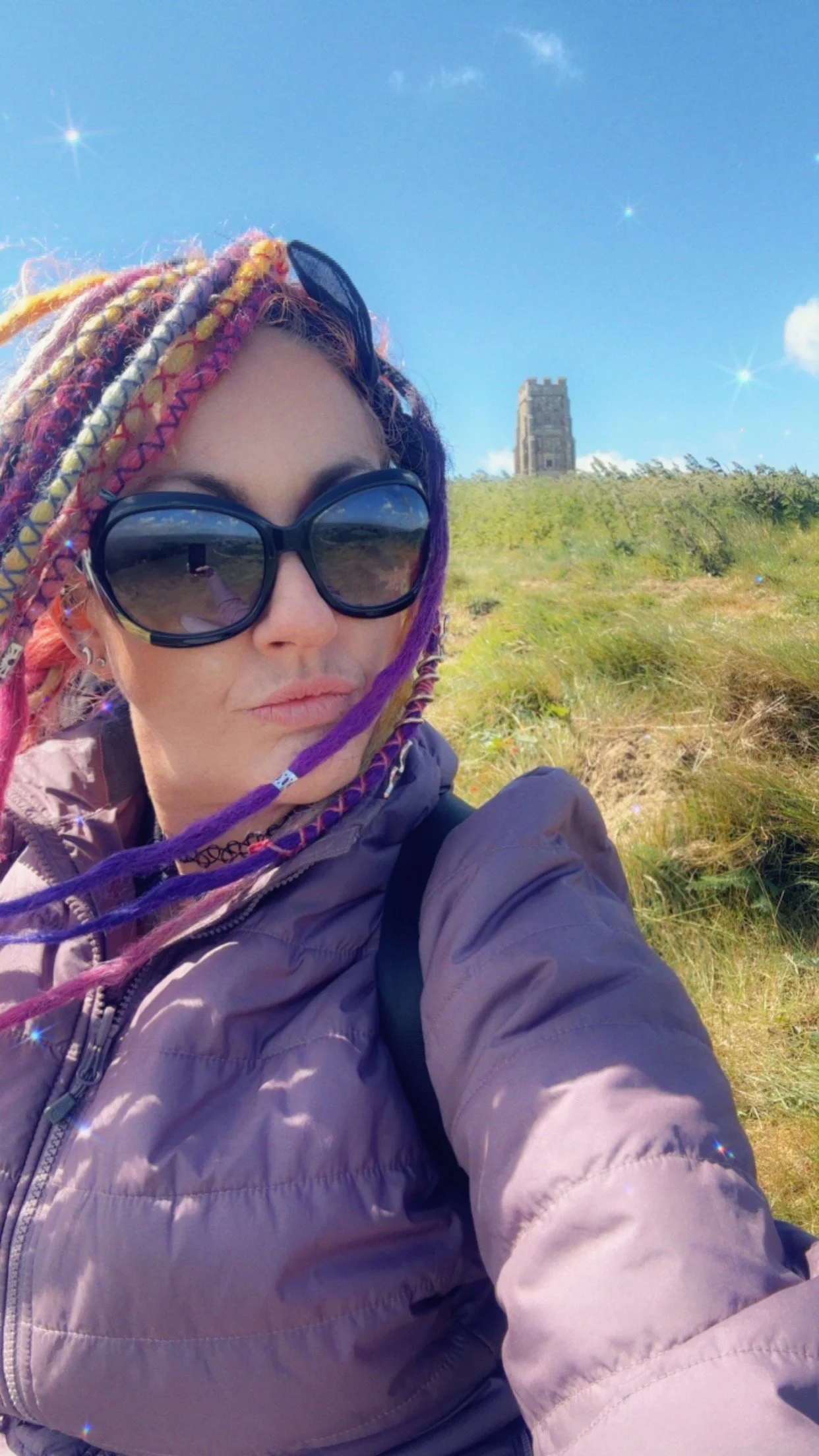 Woman with colorful dreadlocks wearing large sunglasses and a purple jacket taking a selfie outdoors with a hill and an old stone building in the background under a bright blue sky.
