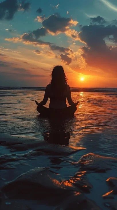 Silhouette of a woman meditating in shallow water at sunset on a beach, with clouds in the sky.