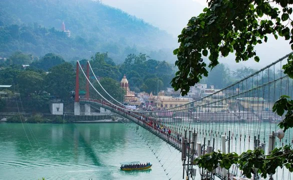 Suspension bridge over a river with boats, surrounded by green hills and buildings in the background.