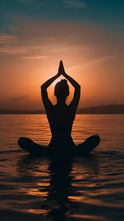 Silhouette of a person practicing yoga in a seated pose with hands in a prayer position, on water during sunset.