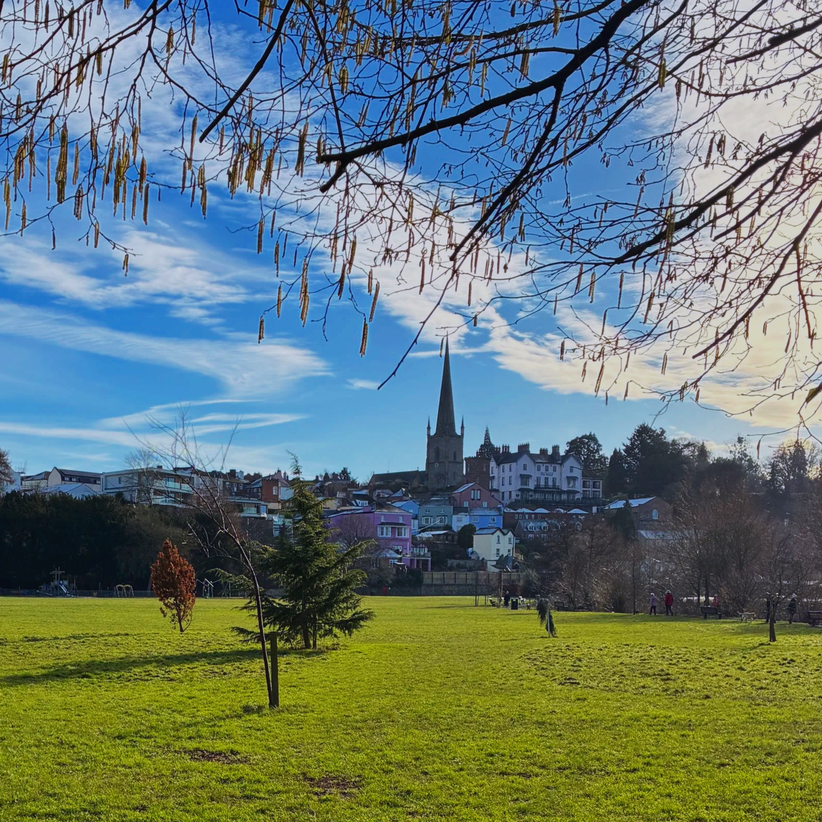 A park with green grass, trees, and people walking or sitting on benches. In the background, there are buildings and a church with a tall steeple under a blue sky with clouds.