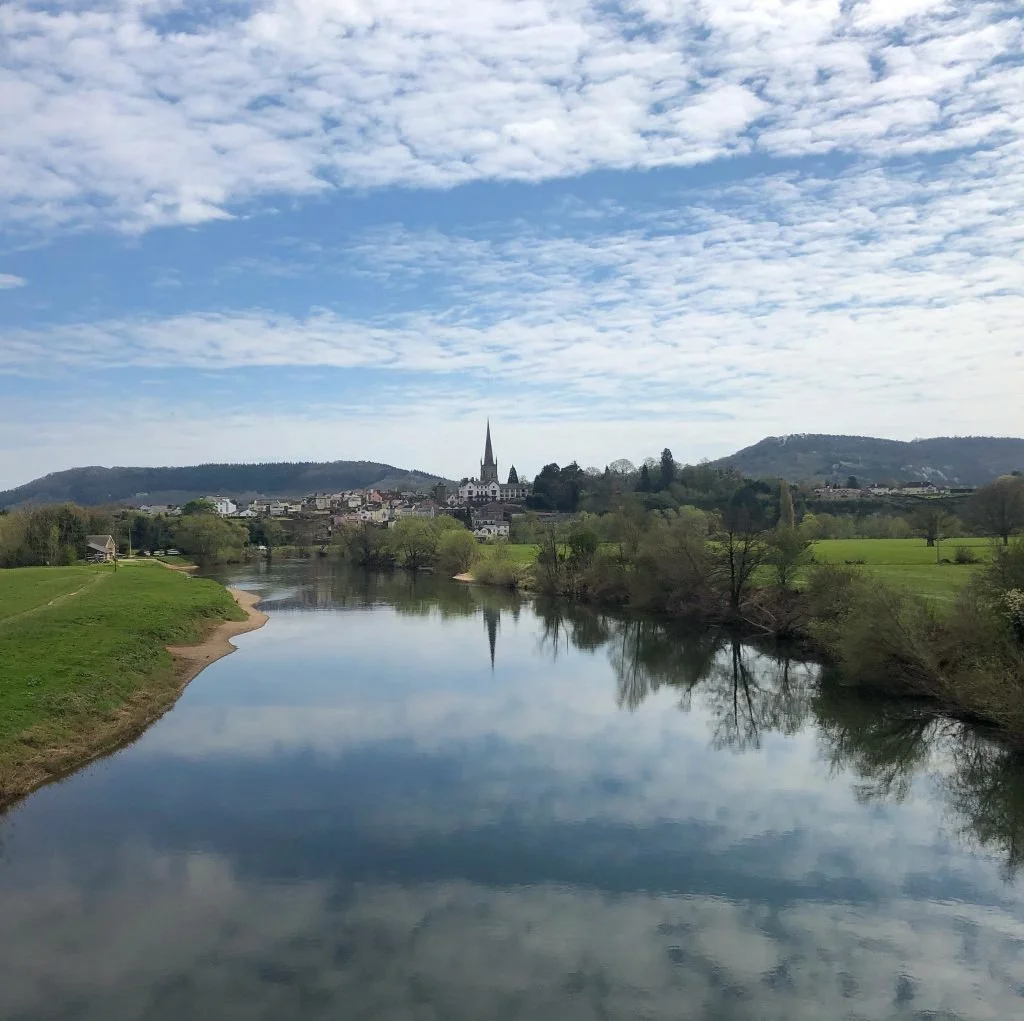 Scenic view of a river reflecting a cloudy sky, with a small town and church spire in the distance, surrounded by green trees and hills.