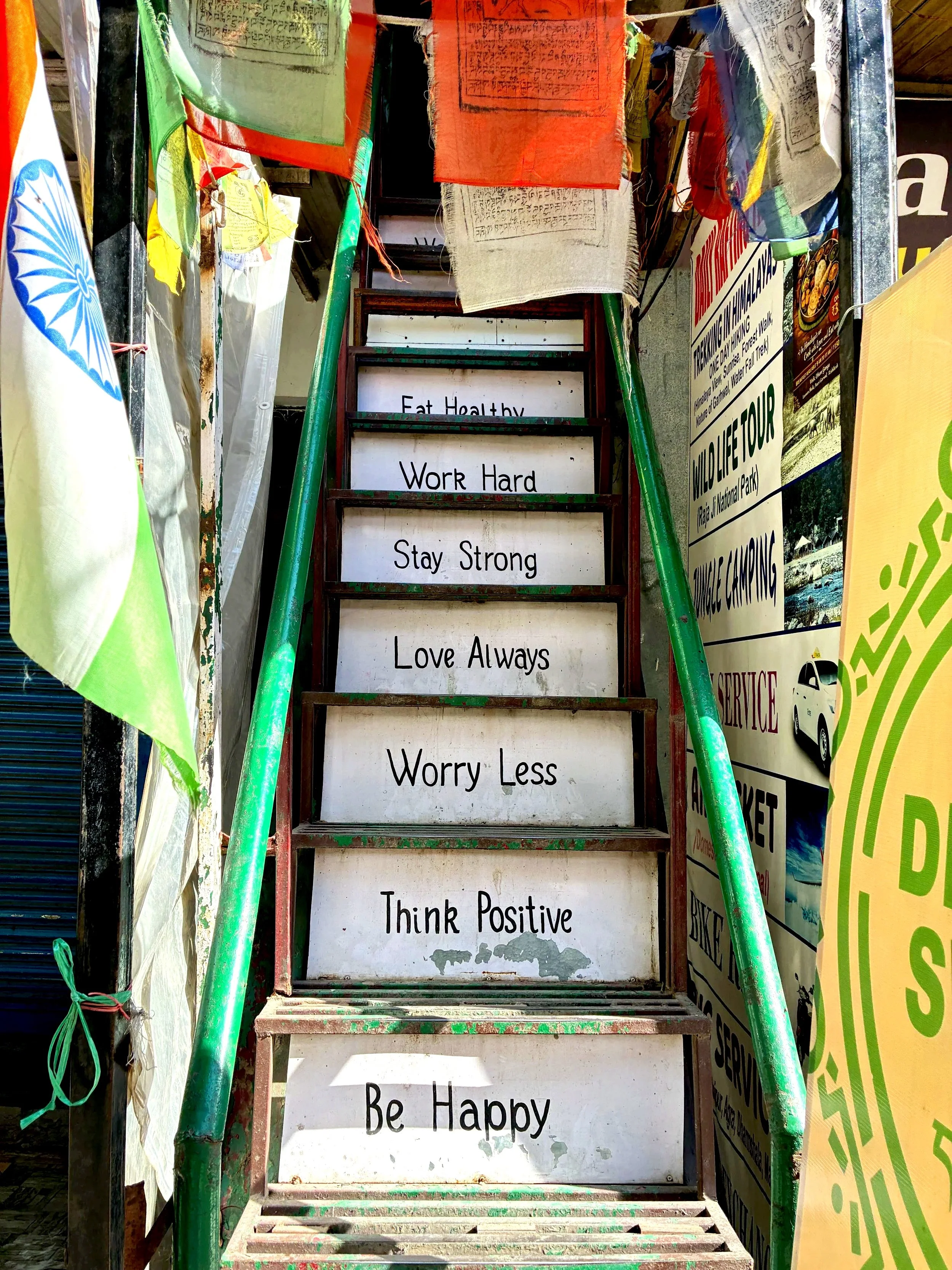 A set of stairs with motivational messages written on each step consisting of: 'Eat Healthy,' 'Work Hard,' 'Stay Strong,' 'Love Always,' 'Worry Less,' and 'Be Happy.' The stairs are surrounded by bright colored flags and posters, suggesting a lively or promotional environment.