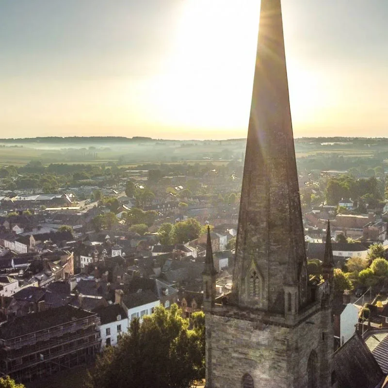 A tall church steeple with a pointed spire standing amidst a small town, with the sun shining brightly in the sky.