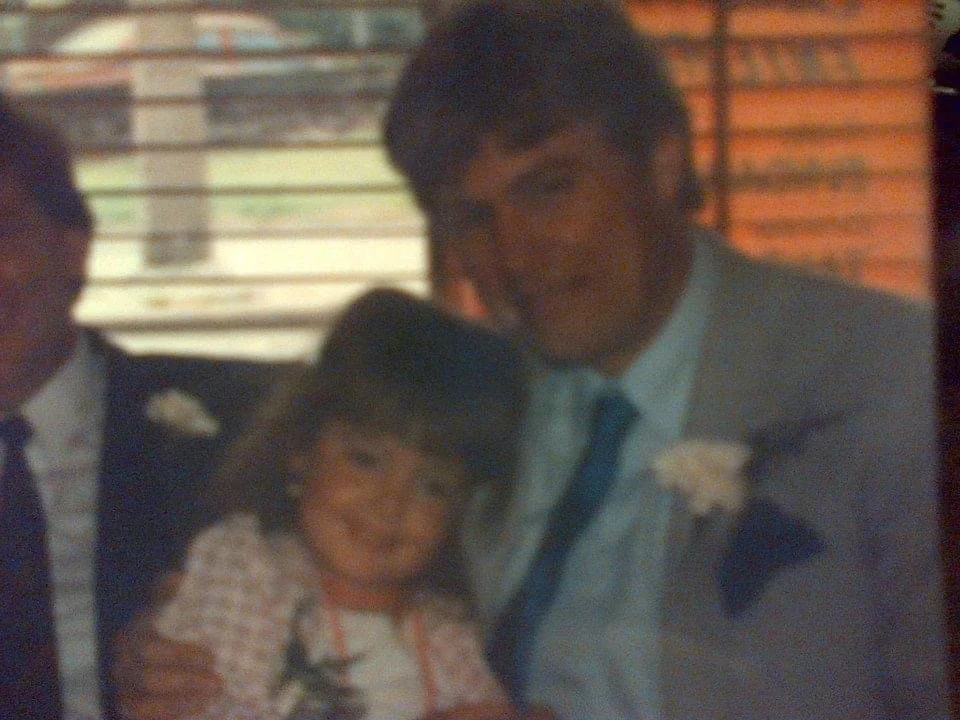 A man in a gray suit and tie with a white boutonniere, a young girl and another man in a suit, all smiling and posing together indoors with a brick wall and window in the background.