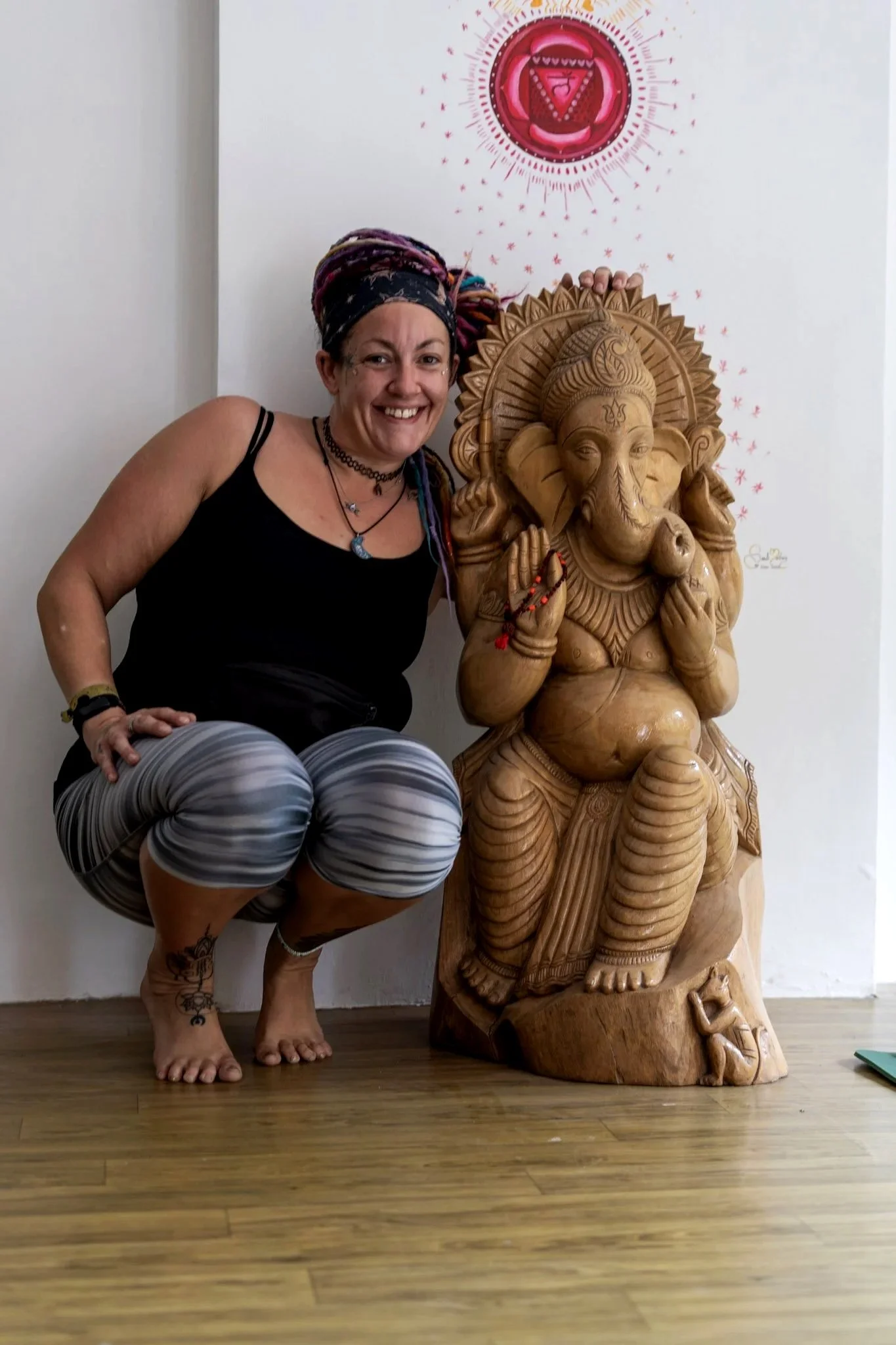 A woman with a colorful head wrap, black tank top, and striped leggings squatting next to a large wooden carved statue of the Hindu deity Ganesha, with religious artwork hanging on the wall behind them.