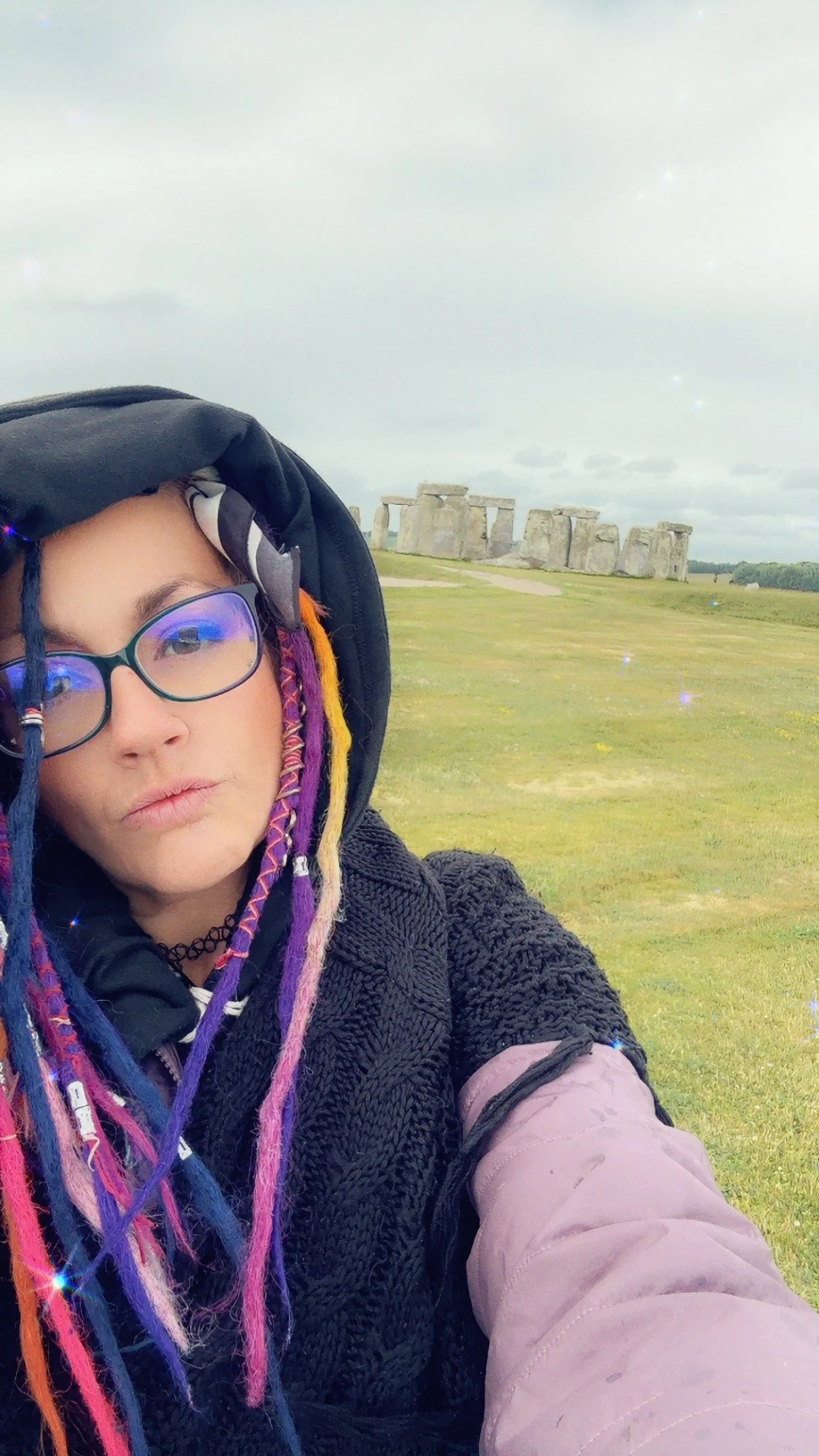 Selfie of a woman with colorful dreadlocks and glasses, wearing a hoodie and jacket, with Stonehenge in the background on a cloudy day.