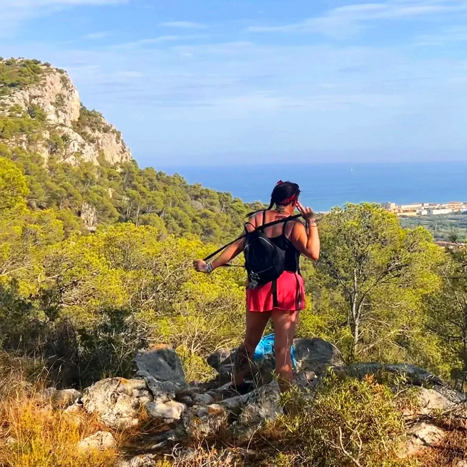 A woman hiking on rocky terrain surrounded by greenery, with a coastal landscape and blue sky in the background.