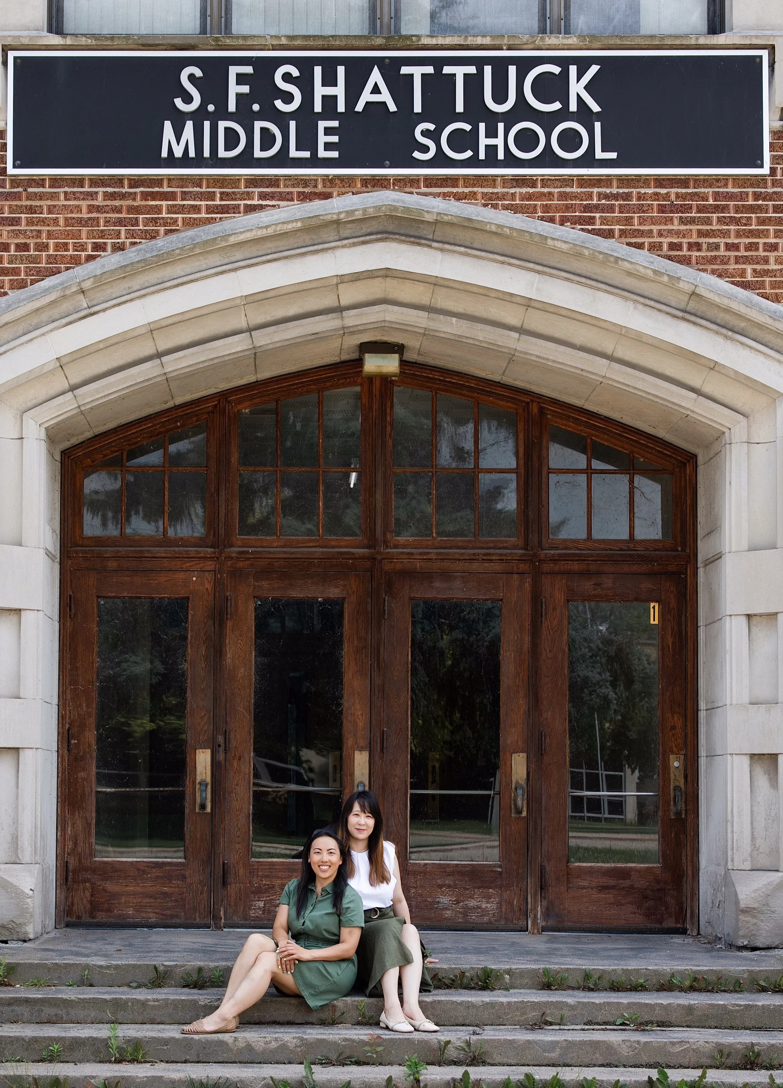 Colleen Bies and Fannie Xie sitting on steps in front of the former S.F. Shattuck Middle School and the current home of the nonprofit they started Fox Valley Community Learning Center, or FVCLC