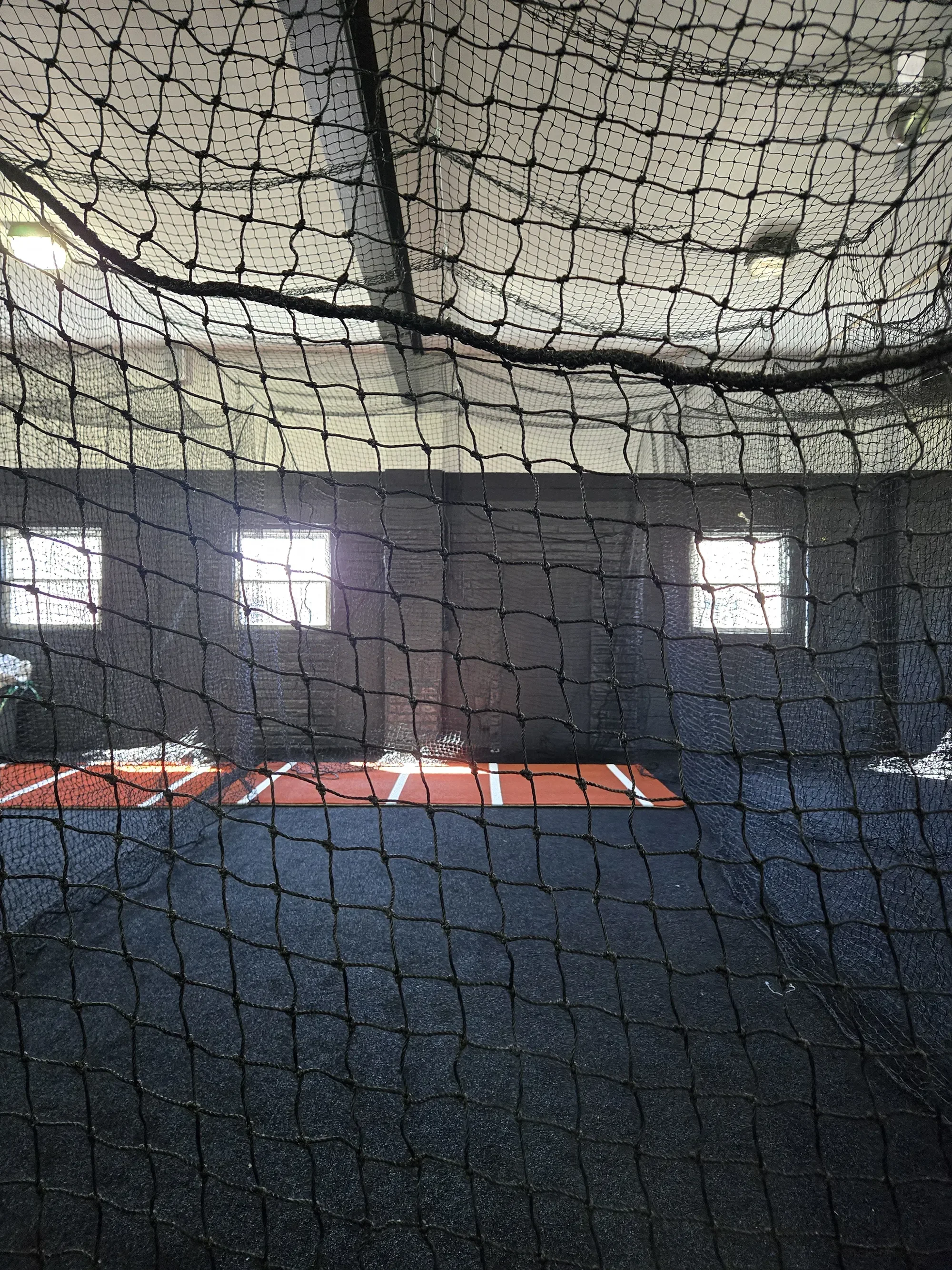 Indoor batting cage with a black net, red and white lines on the floor, and natural light coming through windows.