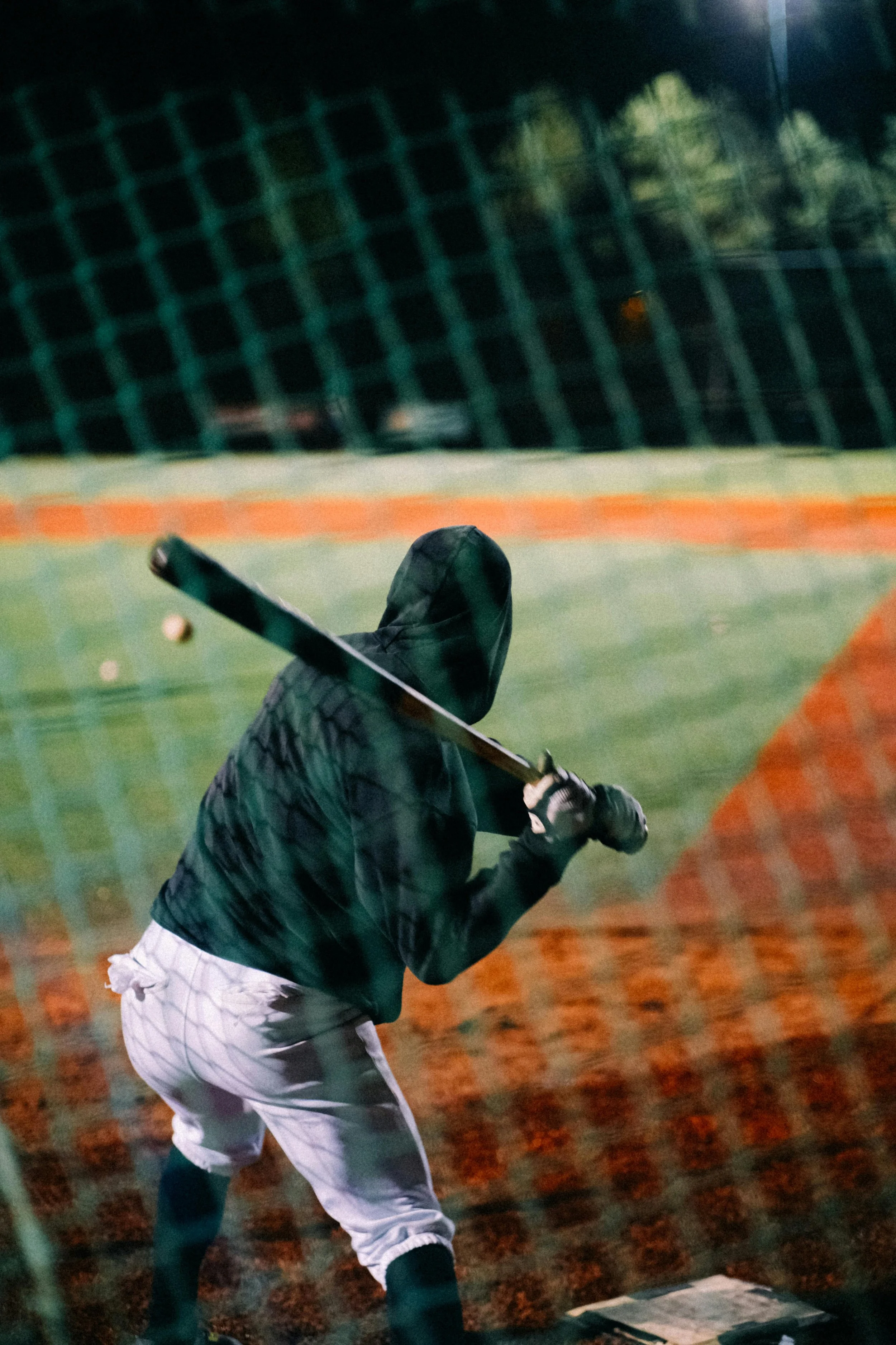 A person wearing a hoodie and white baseball pants practicing batting at a baseball field at night, seen through a green fence.
