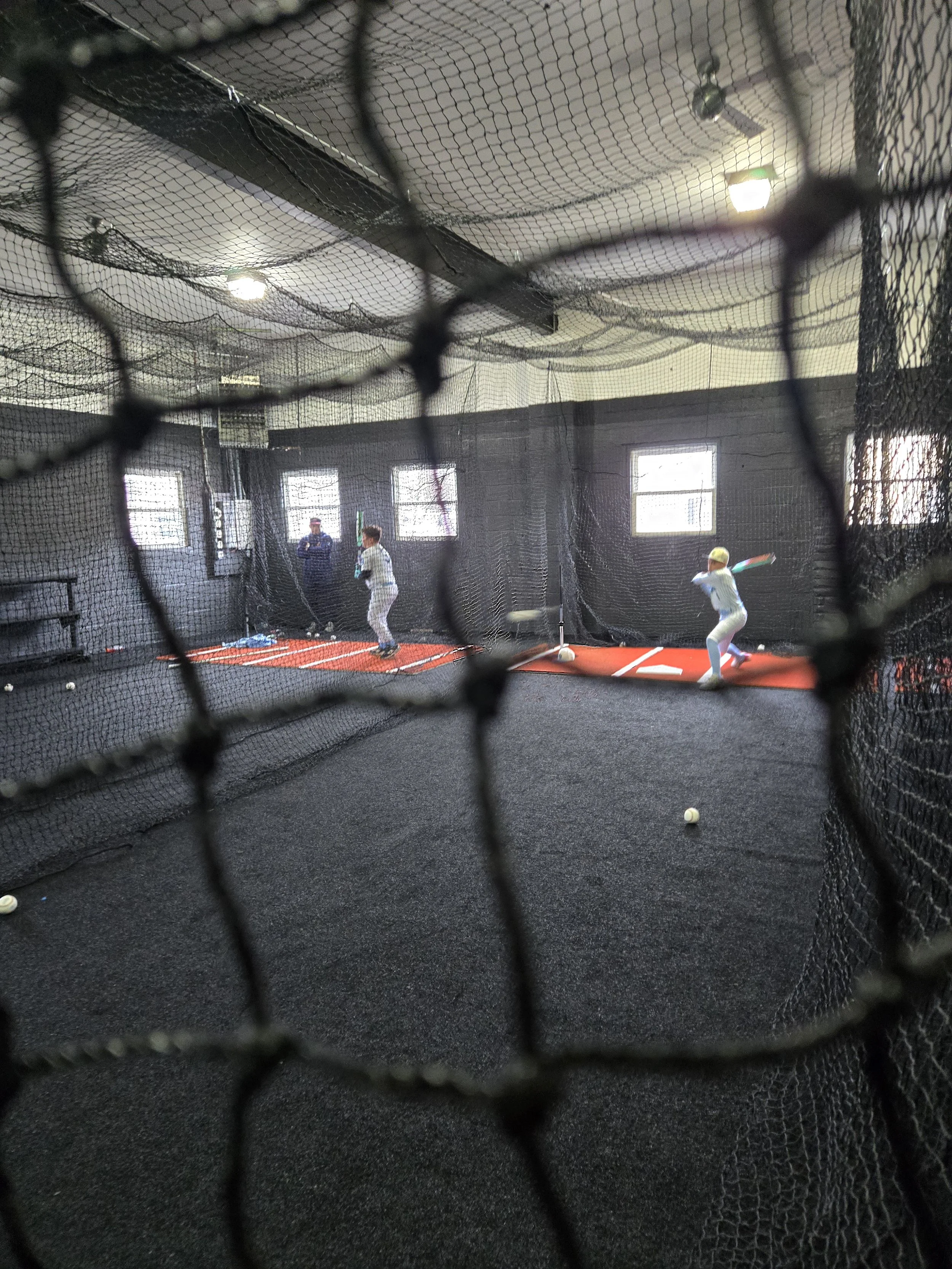 Indoor batting practice area with children swinging baseball bats and a coach, viewed through a safety net.