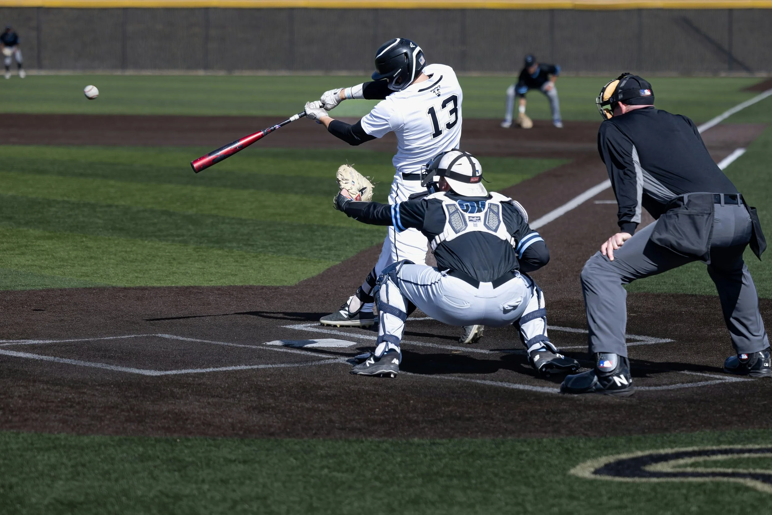 A baseball player wearing a helmet and jersey number 13 is swinging a bat at a pitched ball. The catcher and umpire are positioned behind home plate, with the catcher crouching and the umpire ready to call the pitch. The game is taking place on a baseball field.