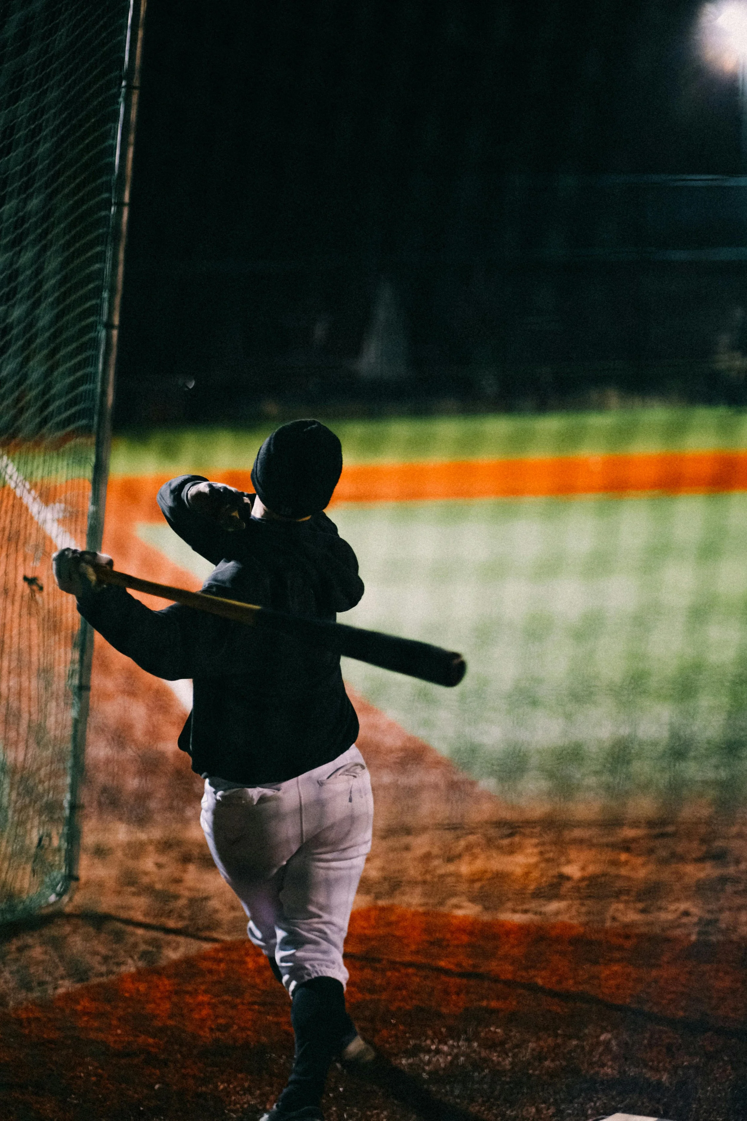 A person playing baseball at night, swinging a bat near a fence, on a well-lit baseball field.
