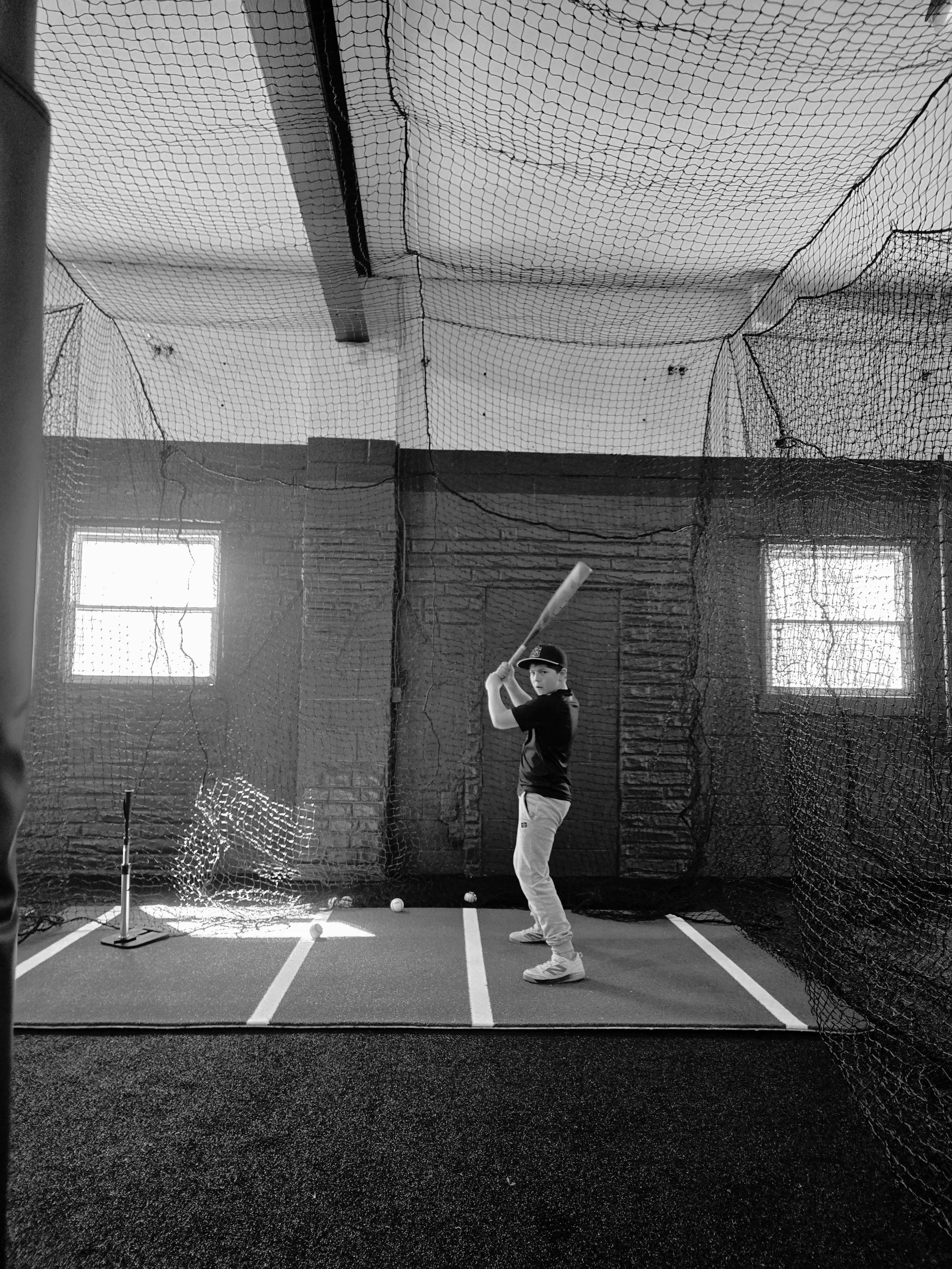 A young boy in a batting stance at an indoor batting cage, holding a baseball bat, with a baseball in front of him on the floor, enclosed by netting.