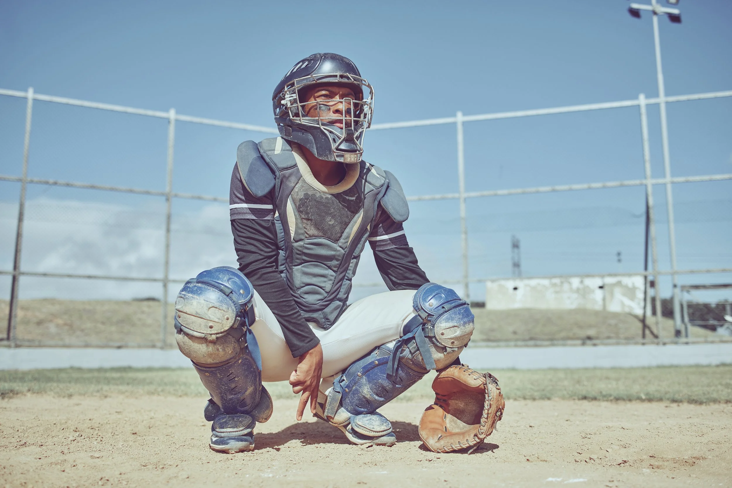 A baseball catcher wearing a helmet, mask, and protective gear crouches on the dirt field, with a baseball glove on his left hand, on a sunny day.