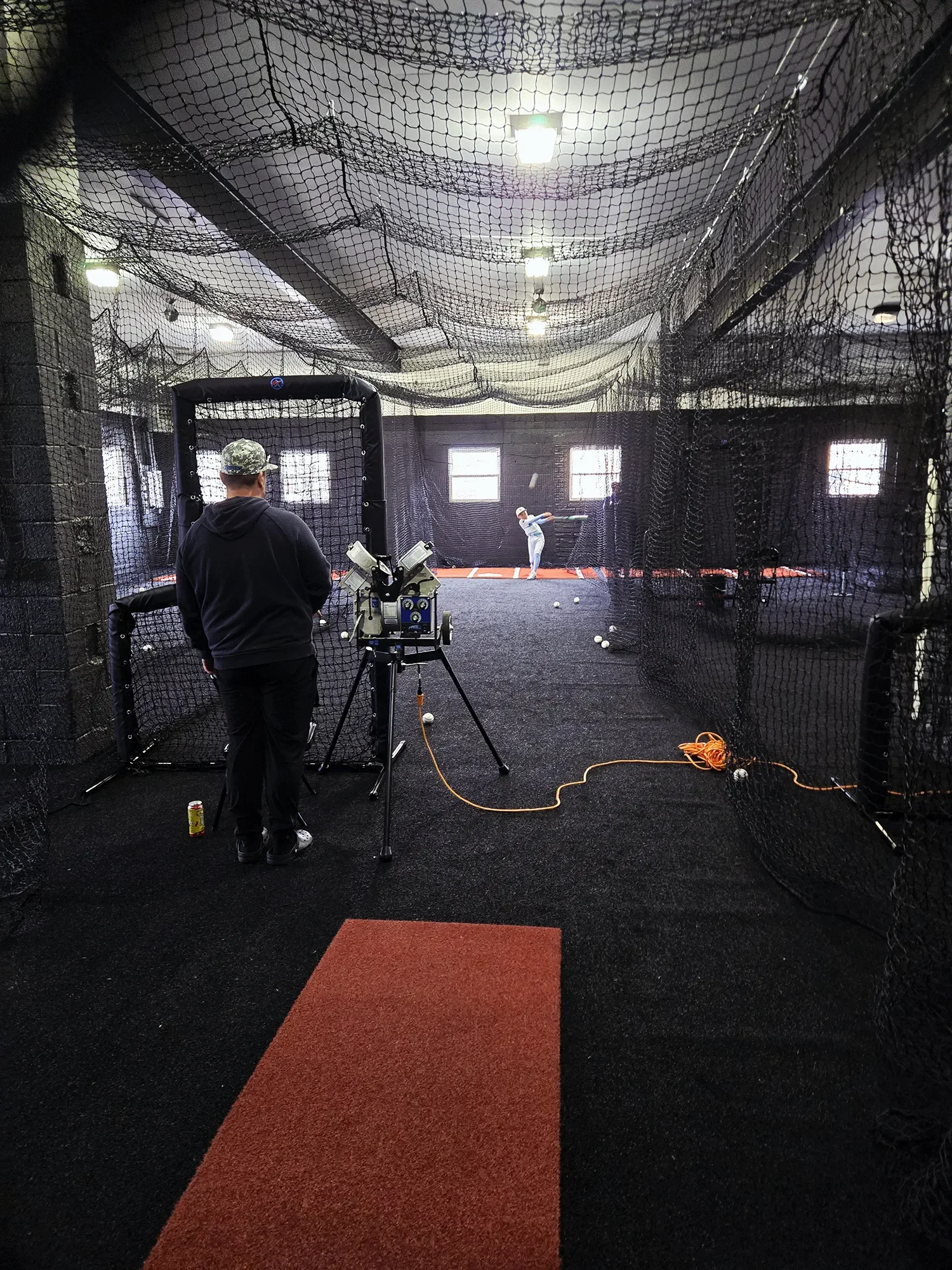 An indoor batting cage with a person practicing baseball swings, a man operating a pitching machine, and a cameraman filming in the foreground.
