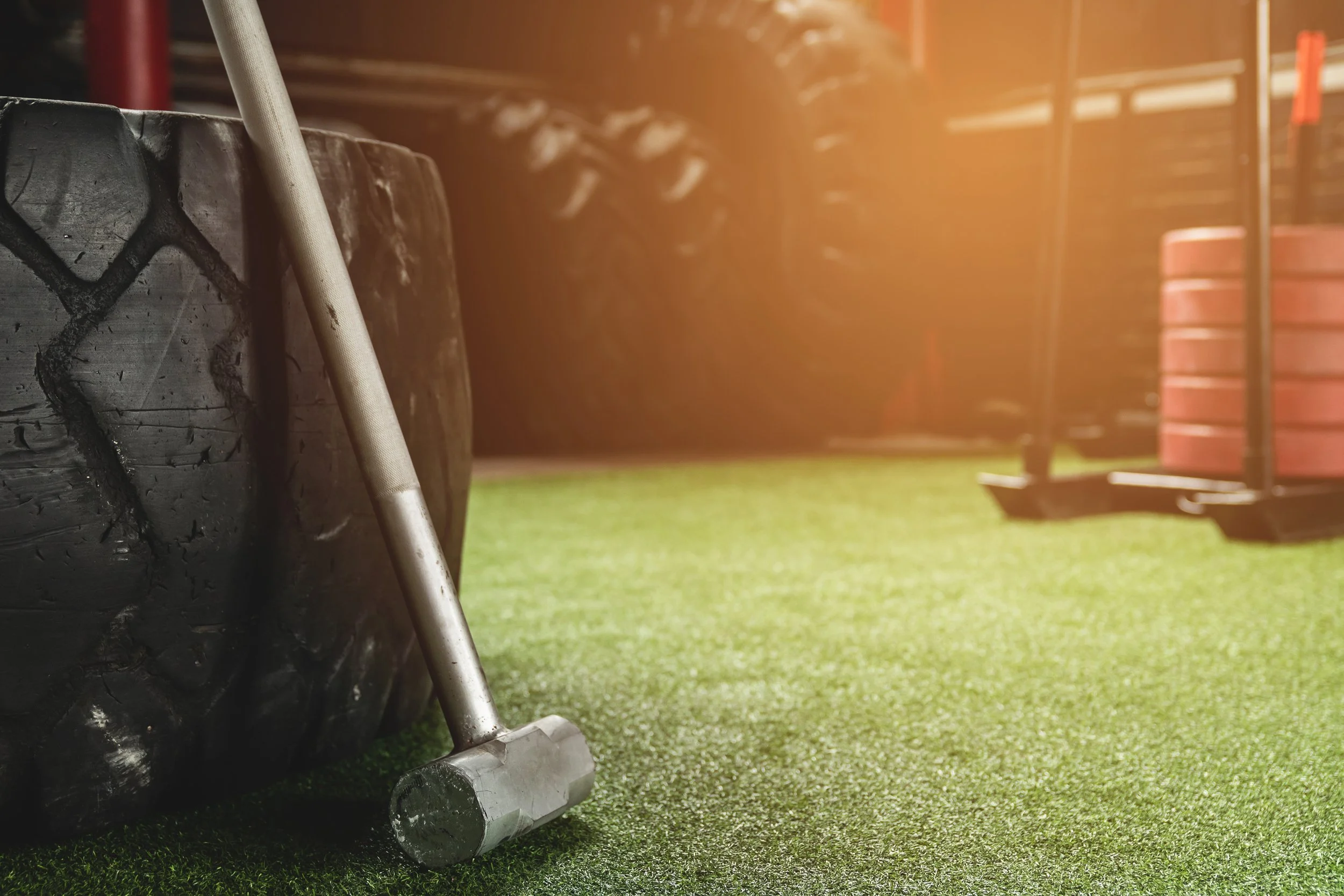 Close-up of a black tire, a metal hammer, and gym equipment on a green artificial turf floor in a gym or strength training area.