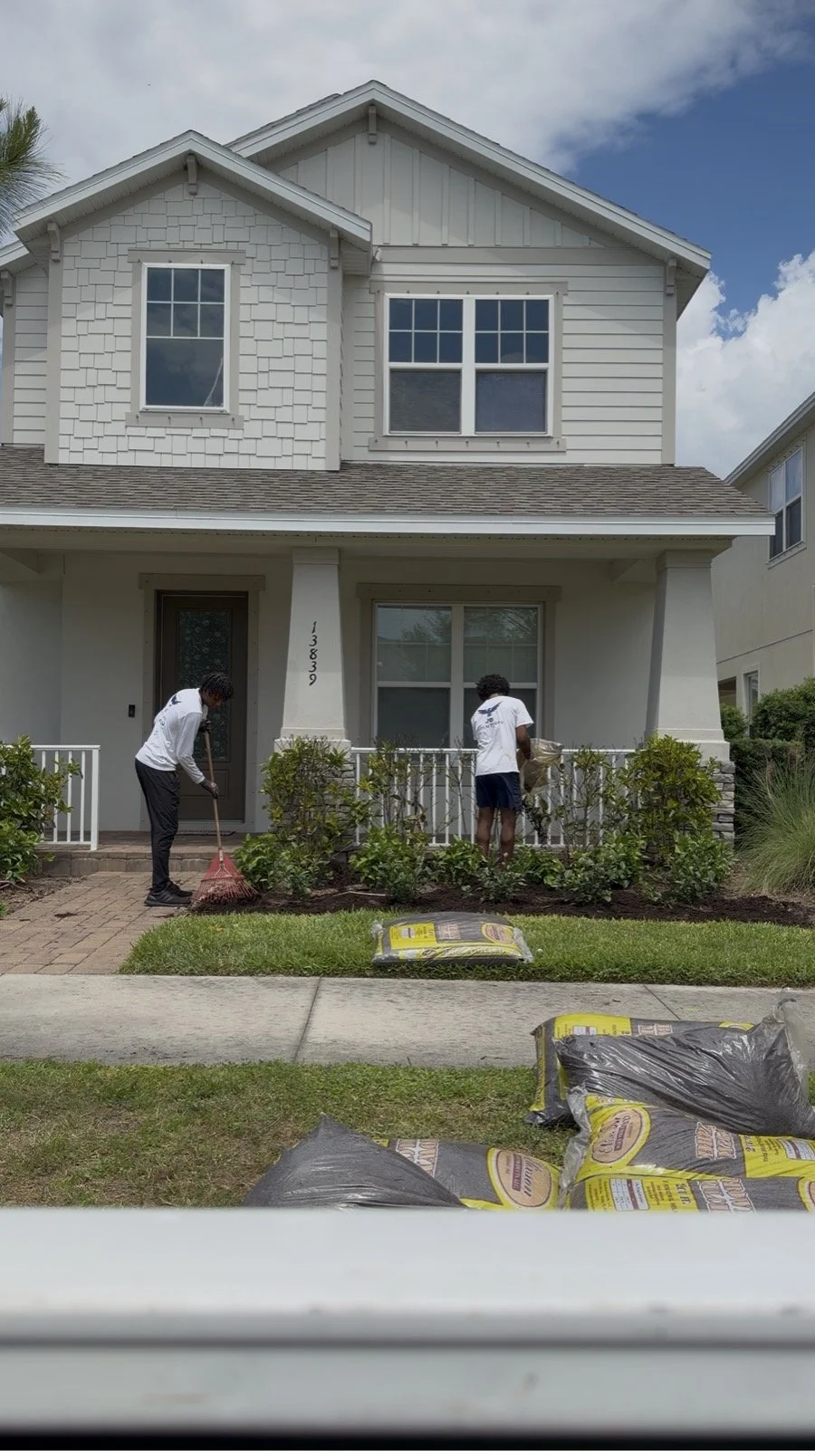 Two young men planting bushes in front of a white two-story house with a gabled roof, with gardening supplies and soil bags in the foreground.