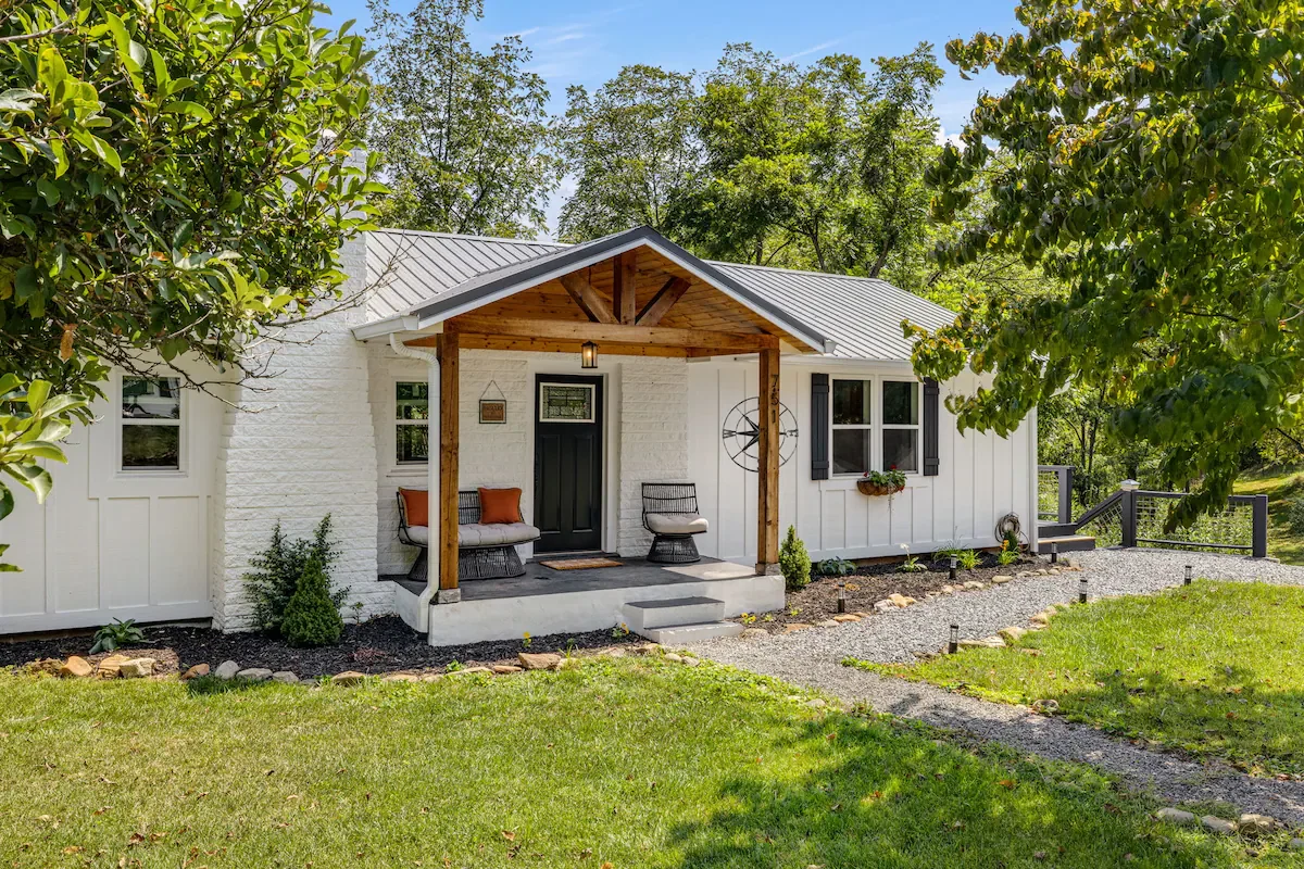White house with a metal roof, small front porch with bench and chairs, surrounded by trees and greenery during daytime.